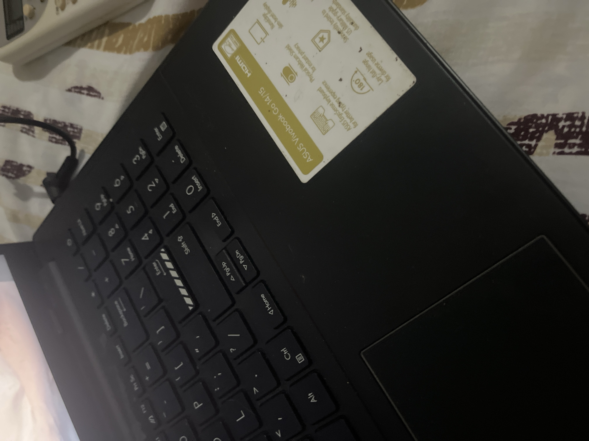 The image shows a close-up, high-angle view of a black ASUS Vivobook Go laptop. The laptop is open, with the keyboard and part of the screen visible. The keyboard has standard QWERTY layout with some additional function keys. A sticker on the right side of the laptop provides product information, including "ASUS Vivobook Go 14/15", "HDMI", and icons representing features like "Nan edge slim-bezel display," "Physical Webcam Shield for instant privacy," "ASUS ErgoSense keyboard for better typing experience," "180° Lay-flat hinge for diverse usage," and "Strengthened to US Military-grade durability standards." A black power cable is plugged into the laptop on the upper right side, and its other end is not visible. The laptop is placed on a surface with a patterned fabric, possibly a bed or couch, with a repeating design of abstract shapes in shades of brown and off-white. A white remote control with buttons is partially visible on the right side of the frame, resting on the patterned fabric. The lighting appears to be dim and ambient. The image does not provide any information that would suggest a specific location within the city of Santa Ana, Venezuela.