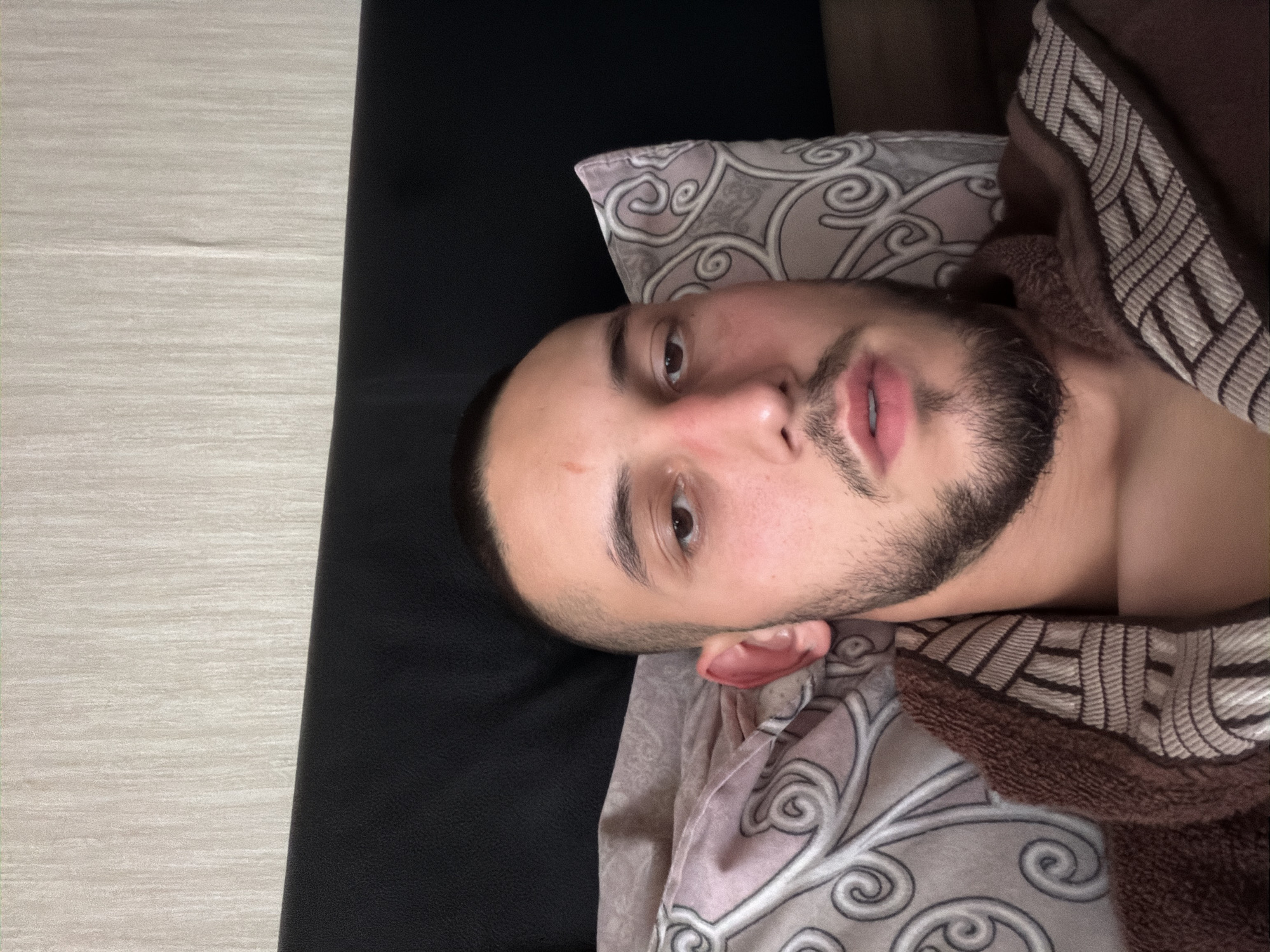 The image displays a young man lying down indoors, captured from a slightly overhead perspective. He appears to be in his 20s or early 30s, with short, dark hair and a neatly trimmed beard. A faint reddish mark is visible on his forehead. He is looking directly at the camera with a neutral, slightly pensive expression, his lips subtly parted.
He rests his head on a light-colored pillow featuring an intricate, grey and white swirling pattern. A brown, patterned blanket or robe, possibly with a soft, textured lining, covers his chest and shoulder. To his left, a dark, plain surface suggests a bed frame or sofa. To his right, a light, textured surface resembling a wooden floor or light-colored rug is visible. The lighting is soft and consistent with an indoor environment. There are no specific visual cues in the image to indicate its location as Sofia, Bulgaria. No text is visible.