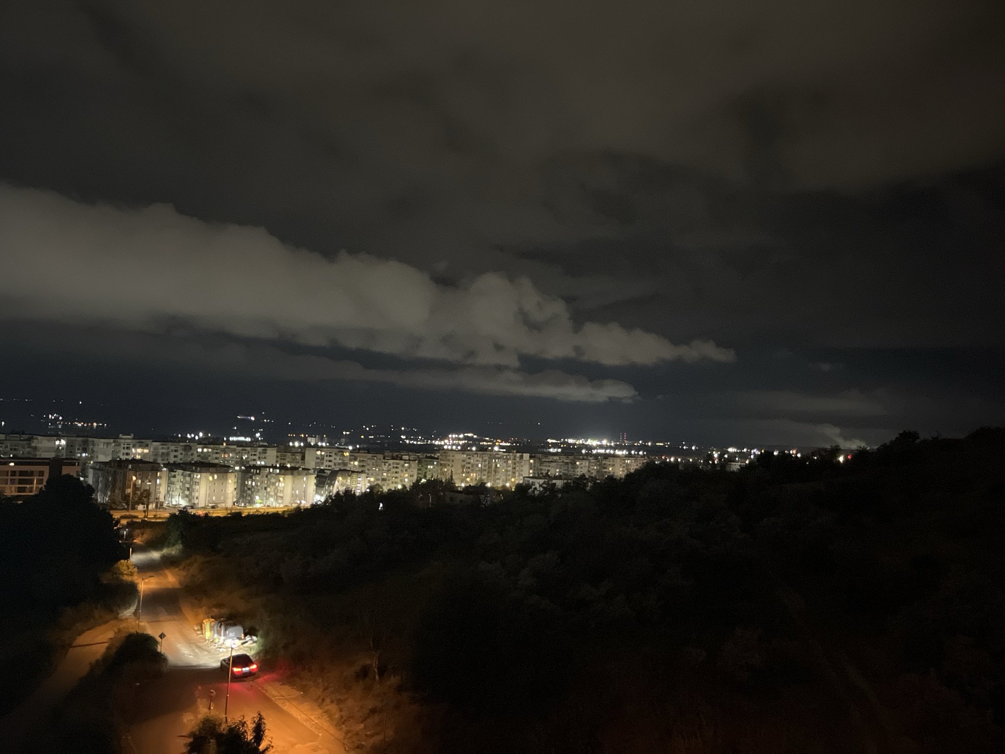 The image captures a night scene overlooking a city skyline, likely Varna, Bulgaria, with apartment buildings illuminated by interior lights. A dark, wooded hillside occupies the foreground and mid-ground, with a winding road cutting through it. A single car with its taillights illuminated is parked on the road, and several large, dark-colored trash bins are visible nearby, with one bin appearing to be overturned. The sky is dark with scattered, illuminated clouds, suggesting a partly cloudy night. The overall impression is one of a quiet, residential area on the outskirts of a city at night.