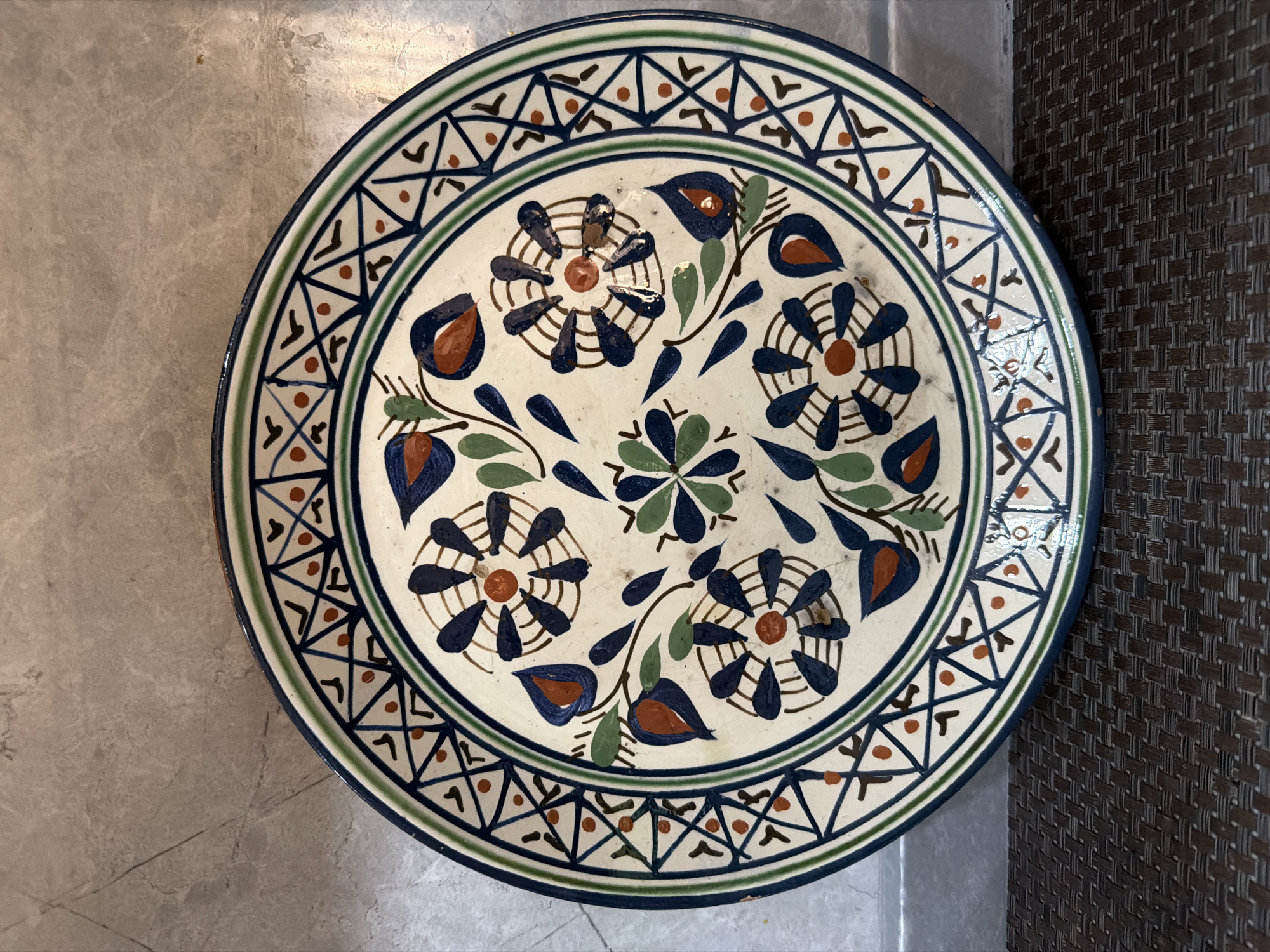 A circular ceramic plate with a floral pattern is displayed against a textured grey wall and a dark woven mat. The plate has a white base decorated with blue, green, and orange motifs. The outer rim features a geometric pattern of triangles and dots in blue and orange. The central part of the plate showcases stylized flowers with blue petals and orange centers, interspersed with green leaves and branches. The plate appears to be an artifact. The image does not provide any information about the city of Sabirabad, Azerbaijan.