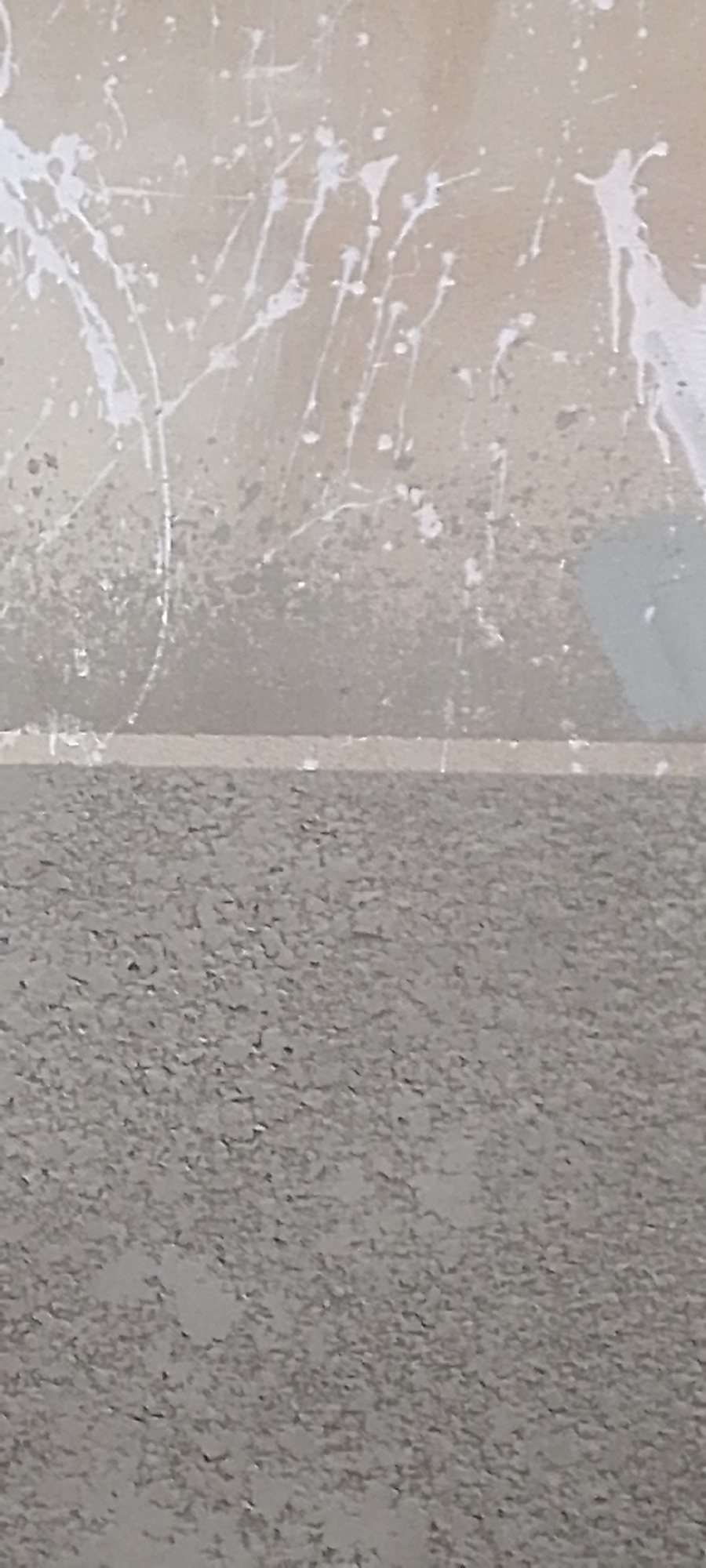 The image shows a close-up view of a textured wall with white paint splatters. The wall appears to be divided horizontally into two sections. The upper section is a lighter, smoother surface with numerous thin, irregular white lines and dots of paint. The lower section is a darker, coarser textured surface, possibly concrete or stucco, with a rough, granular appearance. A thin, horizontal line separates the two sections of the wall. The overall impression is that of an unfinished or weathered surface, possibly an exterior wall. There is no information in the image to suggest its location in Jalingo, Nigeria, or any specific people, subjects, actions, or interactions.