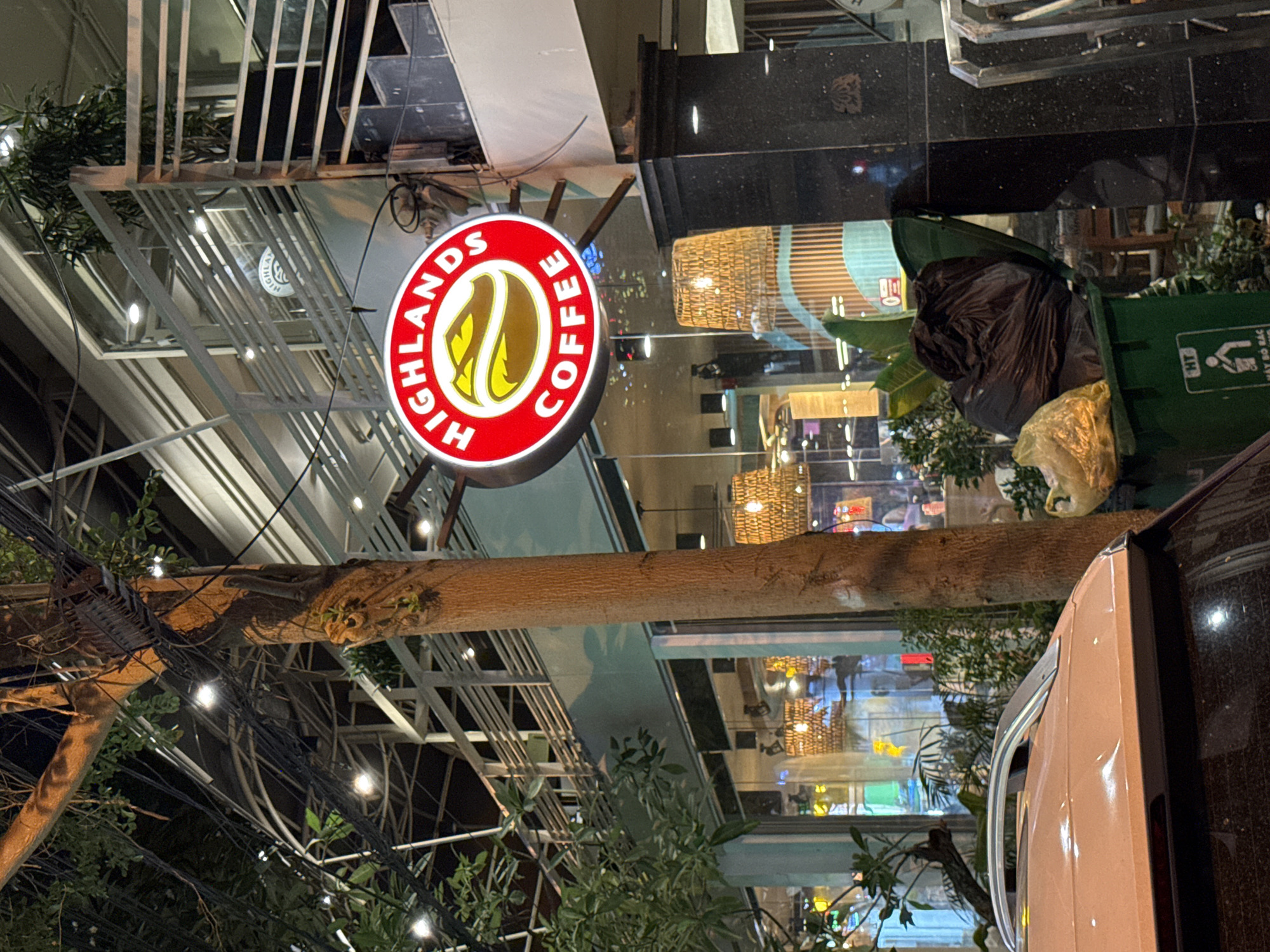 The image depicts the exterior of a Highlands Coffee shop in Nha Trang, Vietnam, at night. A prominent circular red sign with "HIGHLANDS COFFEE" in white lettering and a yellow coffee bean logo is illuminated. The shop has large glass windows revealing interior lights and woven pendant lamps. A tree with visible electrical wires runs vertically in the foreground, obscuring part of the building. A white car is partially visible in the lower left corner, and a green trash bin overflowing with black trash bags and a yellow bag of trash is situated in the lower right, near a black marble pillar. The scene is illuminated by streetlights, shop lights, and decorative string lights.