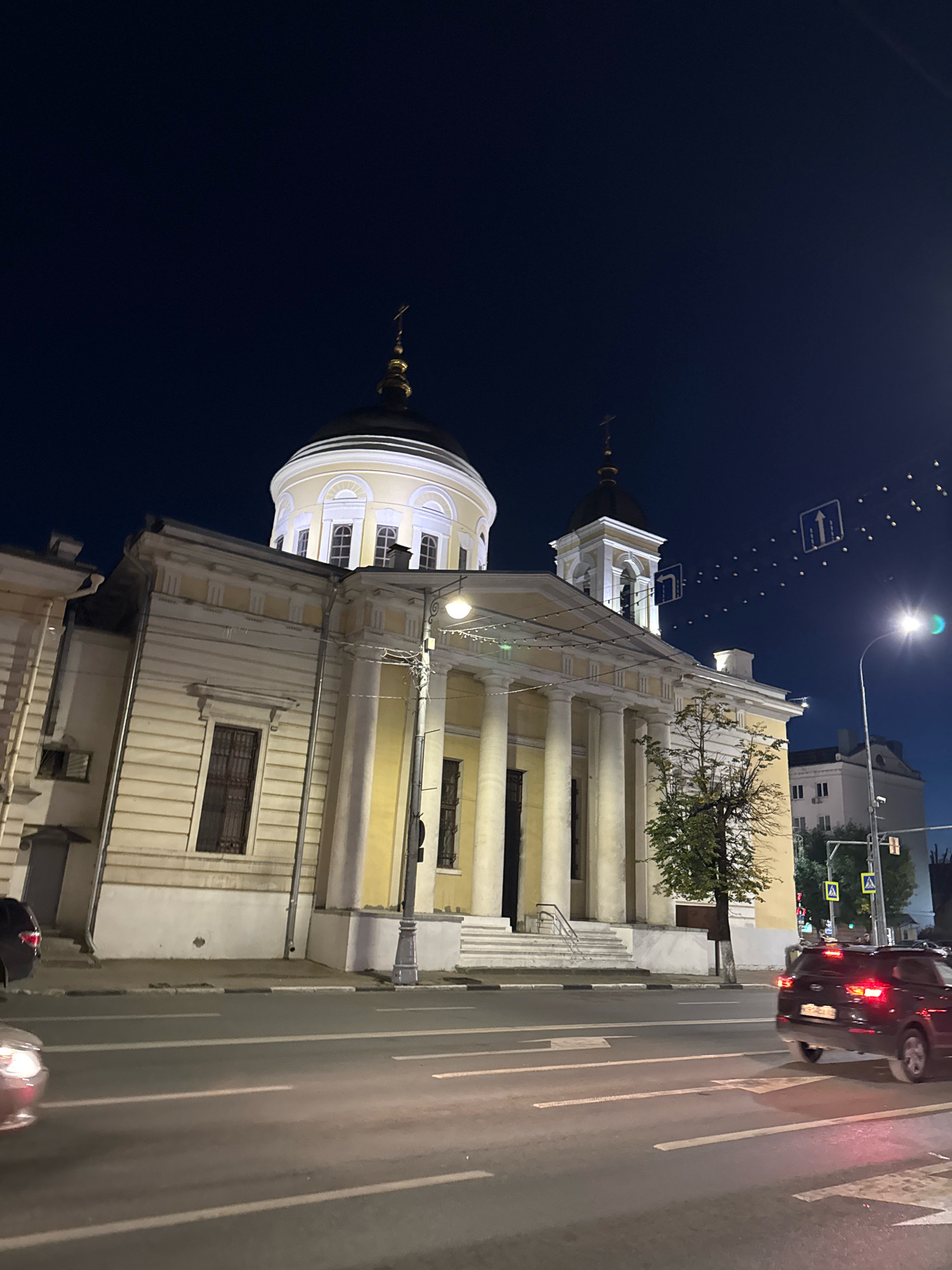 The image shows a night scene in the city of Tver, Russia, featuring a prominent church with a golden dome and a bell tower. The church's facade is yellow and adorned with white columns and decorative elements. A street is visible in the foreground with cars driving on it and streetlights illuminating the scene. Traffic signs are also present, indicating road directions. The sky is dark blue, suggesting nighttime. The overall impression is a calm urban environment with a notable religious building as the focal point.