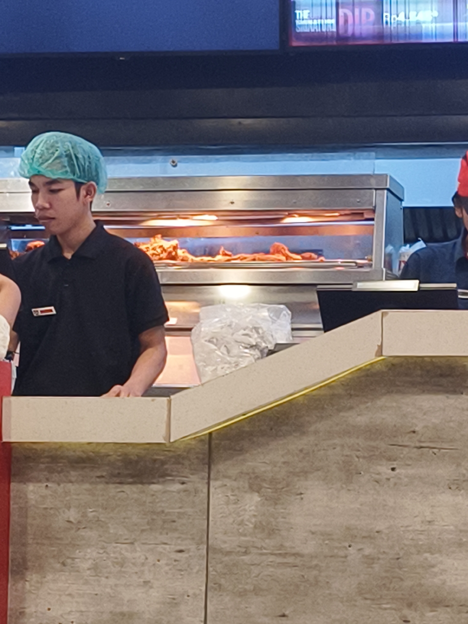 The image shows two male employees behind a counter in what appears to be a fast-food restaurant. The employee on the left is wearing a black polo shirt and a light green hair net, with a nametag attached to his shirt. He is looking down and to his left, with his hands on the counter. Behind him, a food display case is visible, containing cooked food, likely chicken. To the right of the first employee, another employee is partially visible, wearing a dark blue shirt and a red hat. He is looking to his right. The counter is made of a speckled, light-colored material with a yellow accent stripe. The background is dark blue, and a screen displaying text is visible at the top. The scene is likely set in Kuta Selatan, Indonesia, given the context of the request.