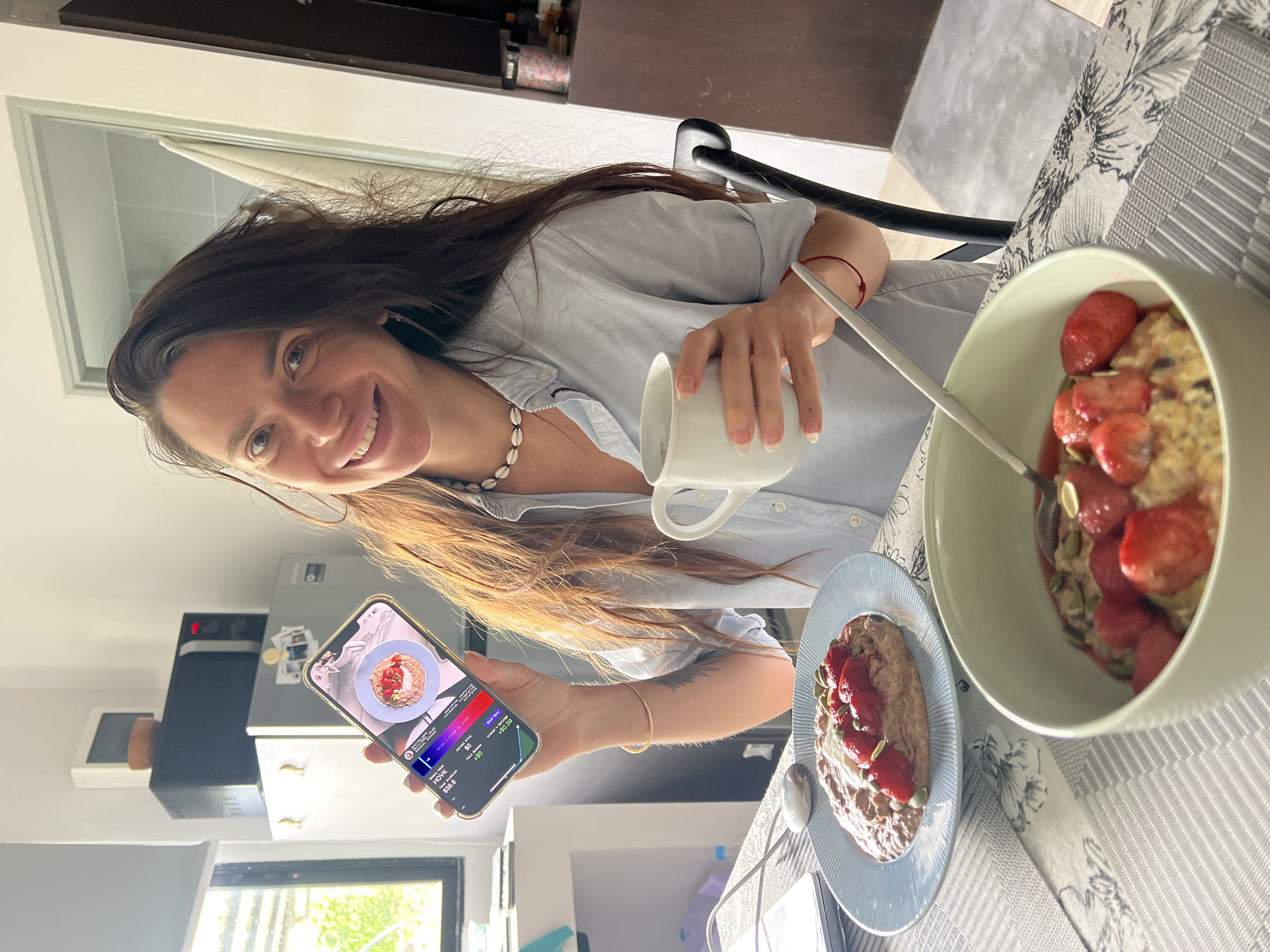 A woman with long, dark hair smiles at the camera while holding a smartphone and a mug of coffee. She is seated at a table with two bowls of oatmeal topped with strawberries and seeds. The phone displays a stock trading app with a graph and figures. In the background, a microwave and refrigerator are visible. The scene appears to be a home kitchen in Ban Rak, Thailand. The woman is wearing a light blue button-down shirt and a shell necklace.