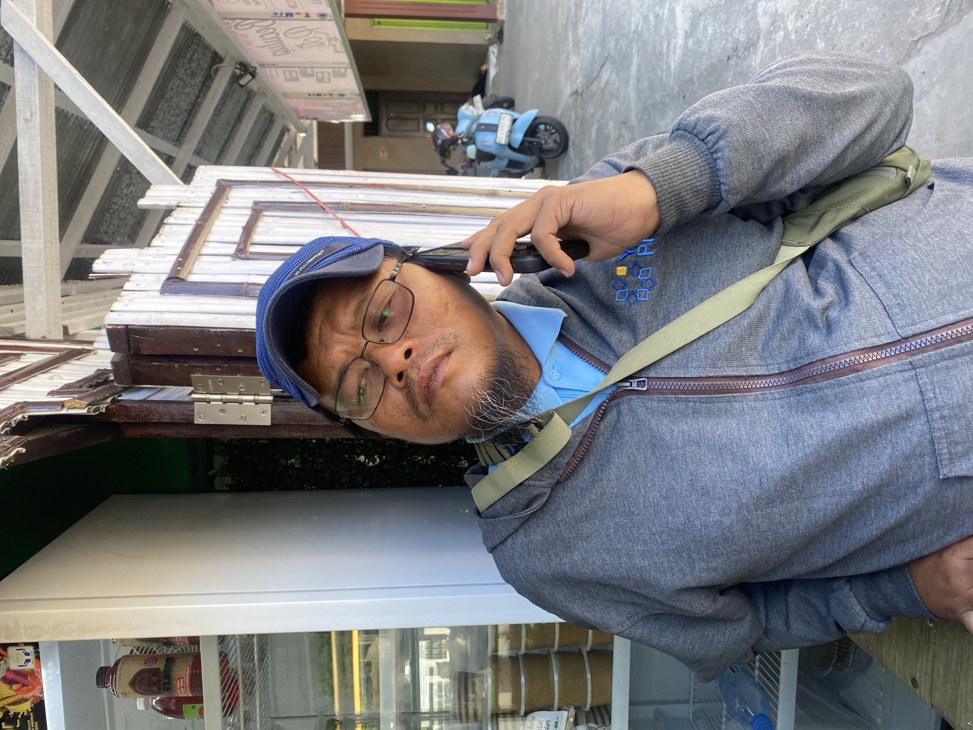 A medium shot captures a man with glasses and a blue cap holding a black smartphone to his right ear. He is wearing a grey zip-up hoodie over a blue polo shirt and a khaki-colored crossbody bag. The man is standing in front of an open doorway with a white wooden doorframe and a weathered, possibly white, paneled wall. Behind him, a glass-fronted refrigerator with various products is visible on the left, and a glimpse of outdoor space with a light blue scooter and what appears to be a person lying on the ground in the background is seen through the doorway on the right. The environment suggests a commercial or street-side setting, possibly in Sungai Paring, Indonesia, with the corrugated metal roof and wooden structures indicating a casual, possibly tropical, location. The lighting suggests it is daytime.