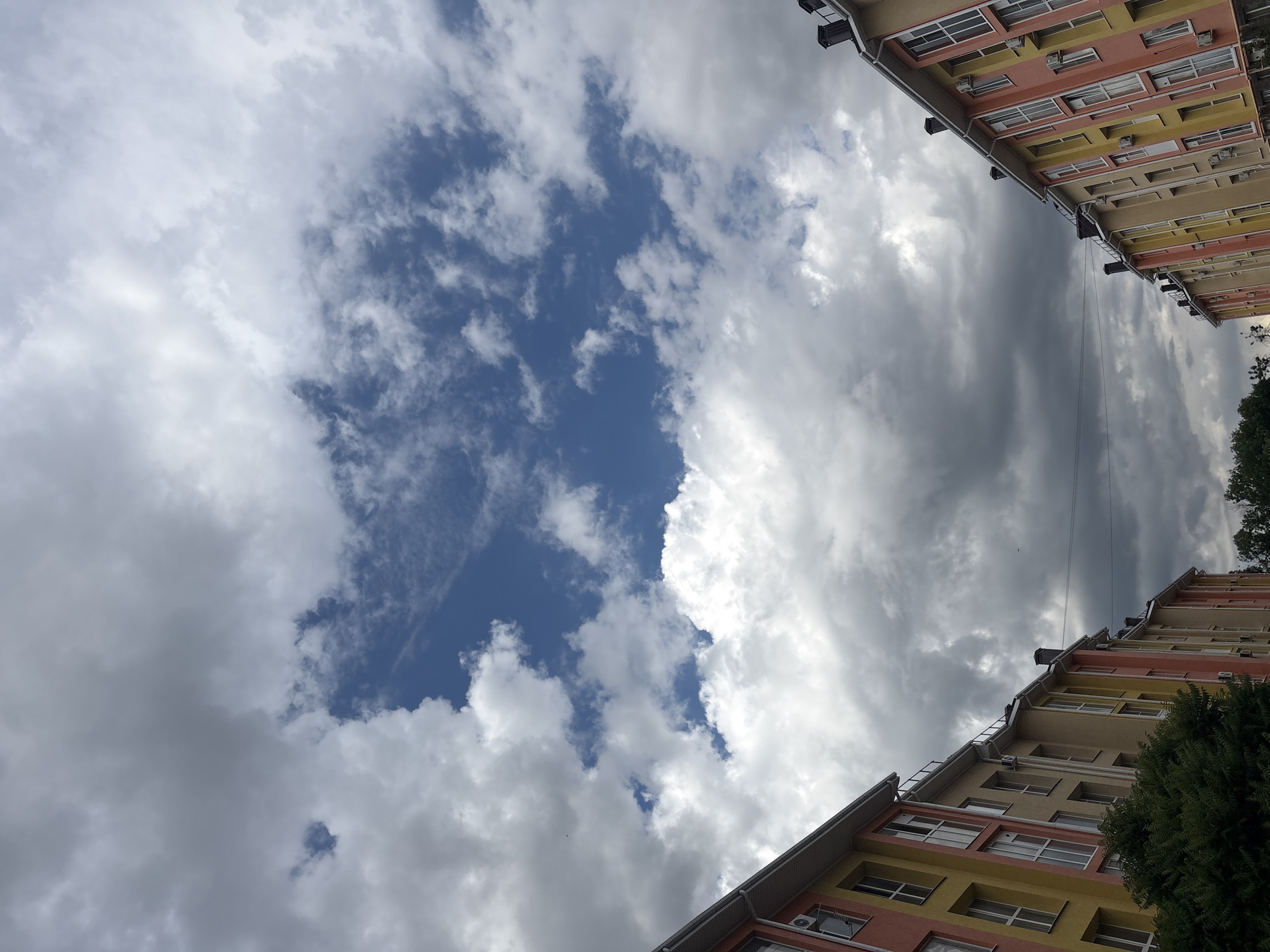 This is a low-angle shot looking up at the sky between two colorful apartment buildings. The buildings are predominantly yellow and pink, with visible windows and air conditioning units. The sky is filled with dramatic clouds, ranging from bright white and fluffy to darker, more ominous gray formations. Patches of blue sky are visible between the clouds. A few thin power lines are strung across the upper part of the image. There is a glimpse of green trees on the right side of the frame, in front of the lower building. The lighting suggests it is daytime, possibly late afternoon or early evening, given the contrast between the bright and dark clouds. The scene depicts an urban environment, with a focus on the sky and the architecture of the buildings. There is no visible text in the image.