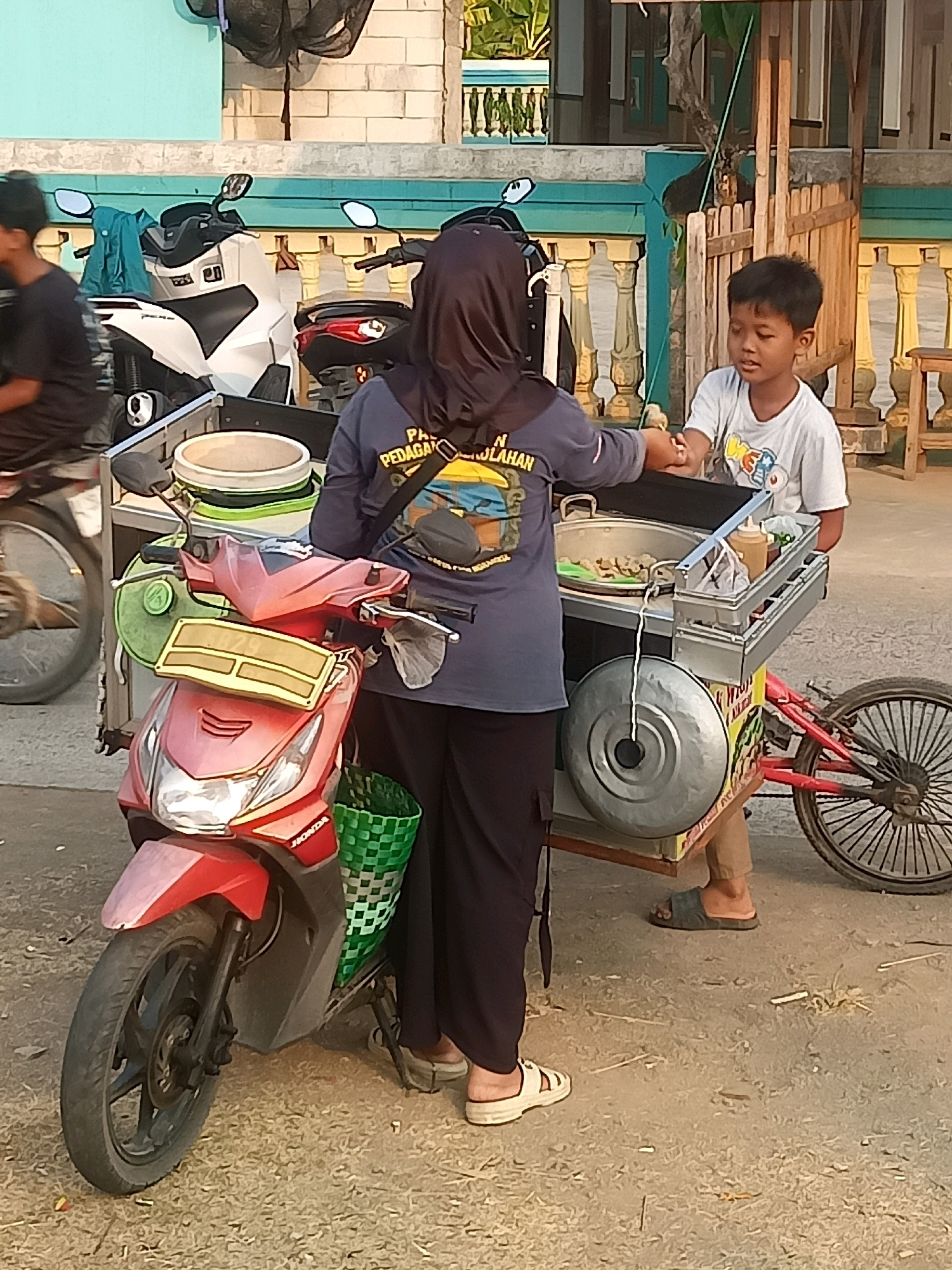 In the city of Sambiduwur, Indonesia, a young boy in a white t-shirt is receiving food from a vendor wearing a dark headscarf and a blue jacket. The vendor is standing behind a food cart, which is attached to a red wheeled frame, and appears to be preparing food in a pot. In front of the vendor is a red scooter. The scene is set outdoors, with a building and a wooden fence visible in the background. The ground is covered with small stones and dirt. The lighting suggests it is daytime, likely late afternoon.
