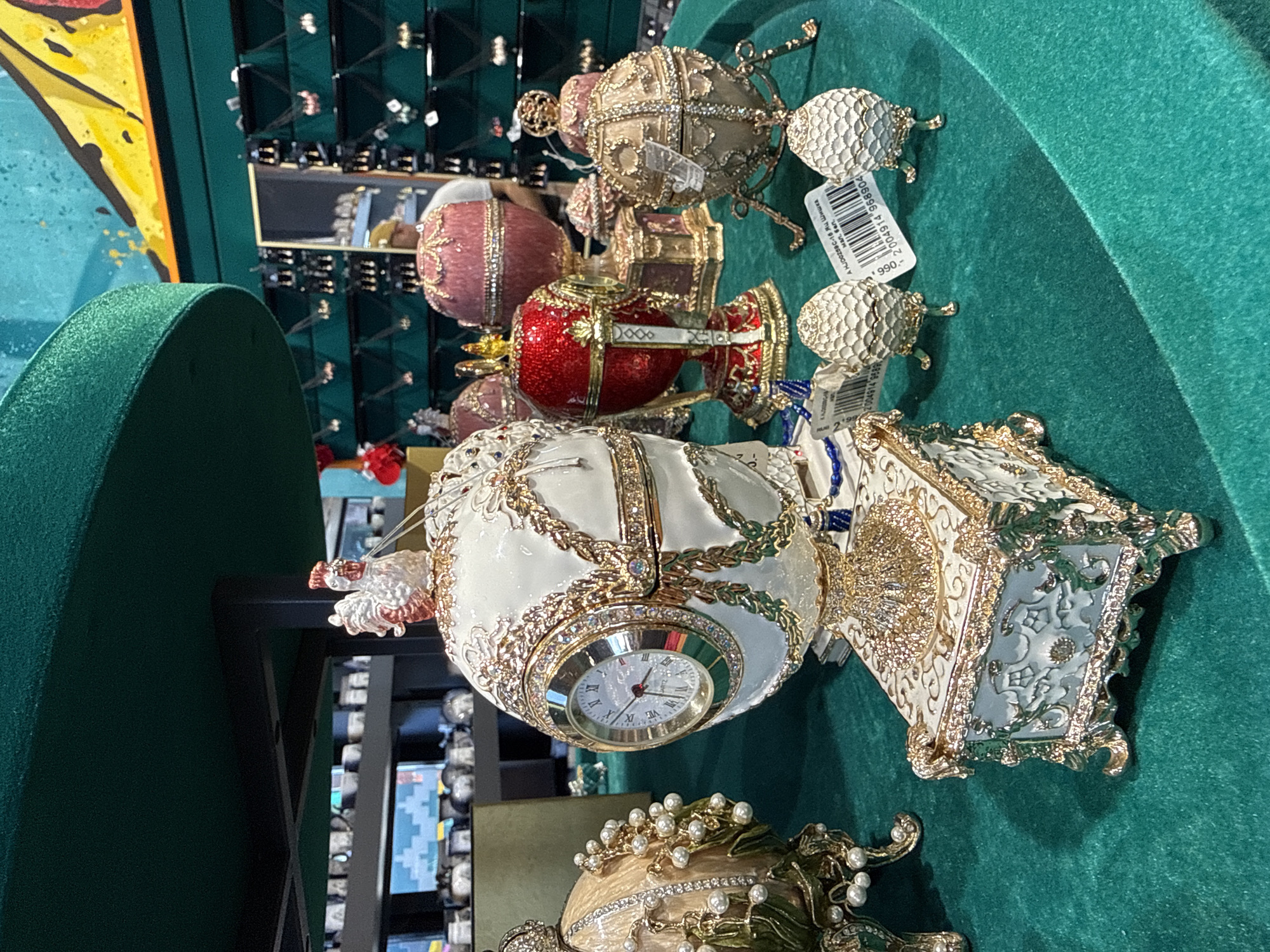 The image depicts a display of ornate, Faberge-style eggs and decorative boxes on a green velvet surface, likely in a shop in Kronstadt, Russia. In the foreground, a large white egg with gold filigree and a clock face is prominently featured, resting on a decorative white and gold box. To its left is a beige egg adorned with pearl-like beads and green leafy embellishments. Behind these, several other egg-shaped decorative items in red, pink, and gold are visible, some on stands. Two smaller white egg-shaped objects with gold stands are in the lower right portion of the image, each with a price tag. In the background, shelves lined with various trinkets and jewelry are dimly visible, and a person wearing a cap is partially seen in the reflection of a mirror. The overall setting suggests a retail environment specializing in decorative gifts.