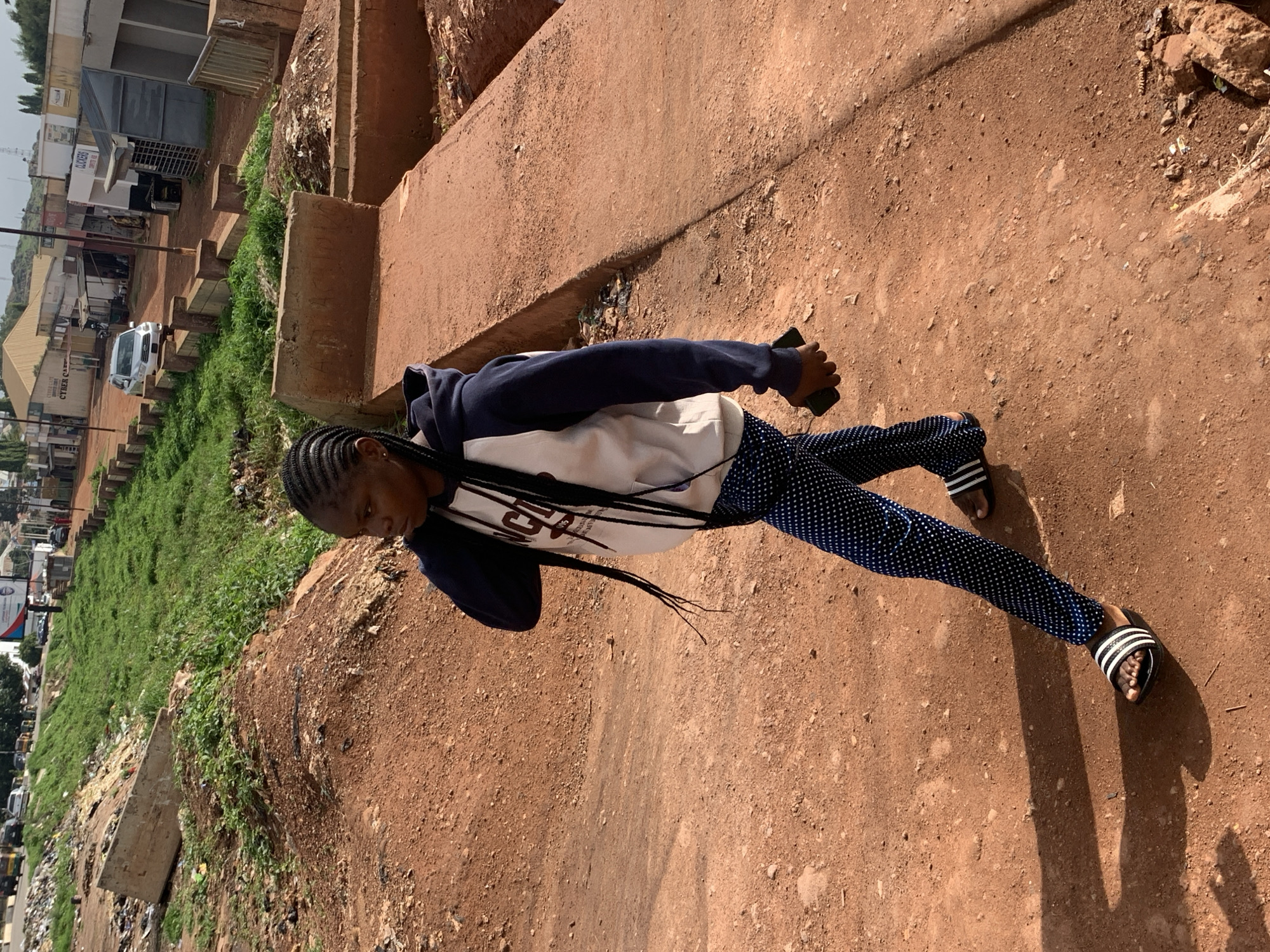 The image depicts a young Black woman walking on a dirt path in what appears to be an urban area in Gura Top, Nigeria. She has braided hair extending past her shoulders and is wearing a white hoodie with dark sleeves and blue polka dot pants. Her footwear consists of black slides with white stripes. She is holding a dark-colored mobile phone in her right hand. The environment is characterized by red dirt, sparse vegetation, and concrete structures, including what looks like a retaining wall or embankment. In the background, there are buildings, a road with vehicles, and more greenery. The overall impression is one of a somewhat undeveloped or developing urban setting.