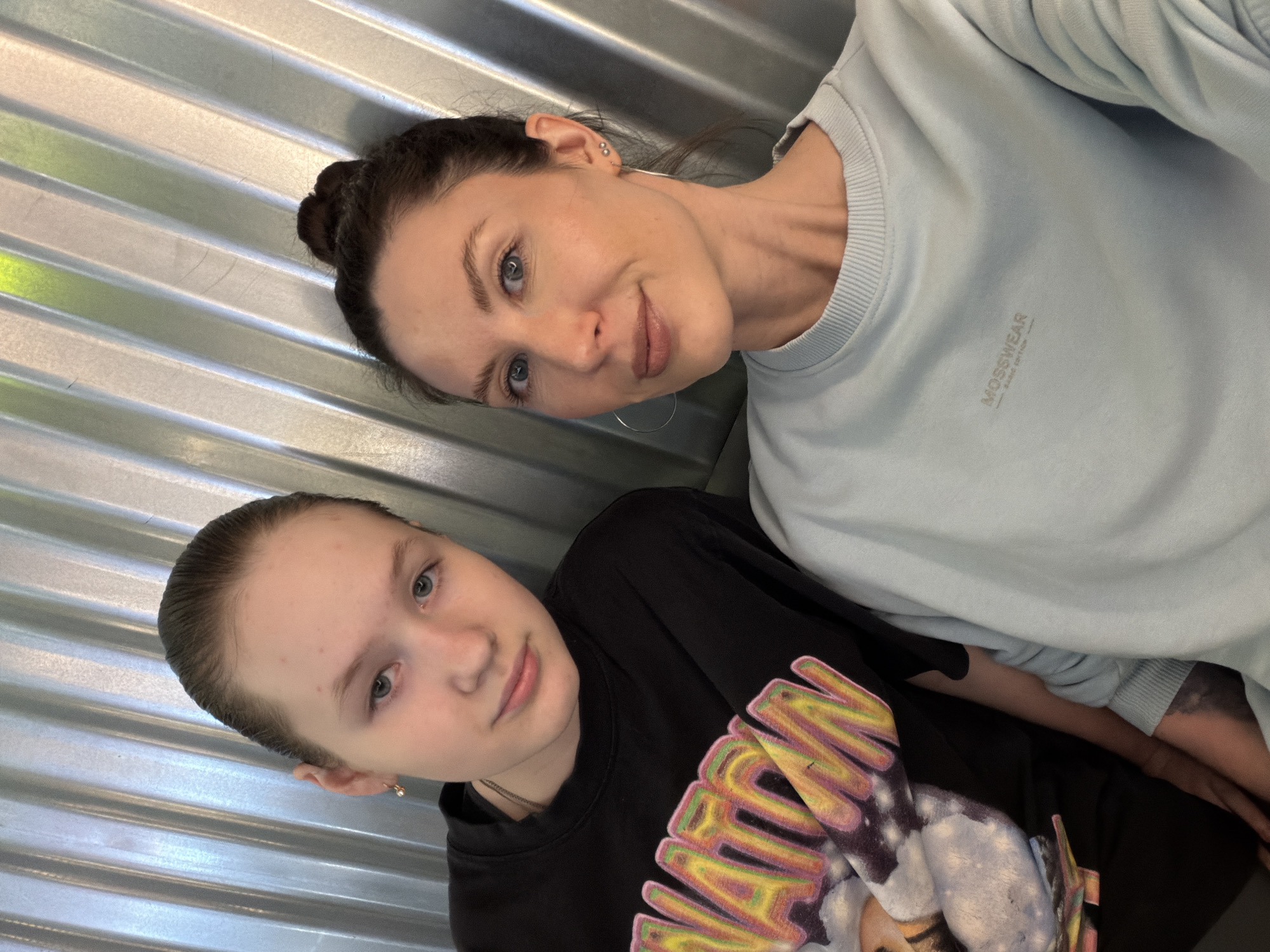 Two Caucasian females pose in front of a corrugated metal wall. The first female, positioned lower in the frame, wears a black sweatshirt with colorful graphic designs. The second female, situated above and slightly to the right, wears a light blue sweatshirt with the word "MOSSWEAR" printed on the upper left chest. Both individuals look directly at the camera with slight smiles.
Location: Perm, Russia (based on the presence of the "MOSSWEAR" brand, a brand originated in Perm).