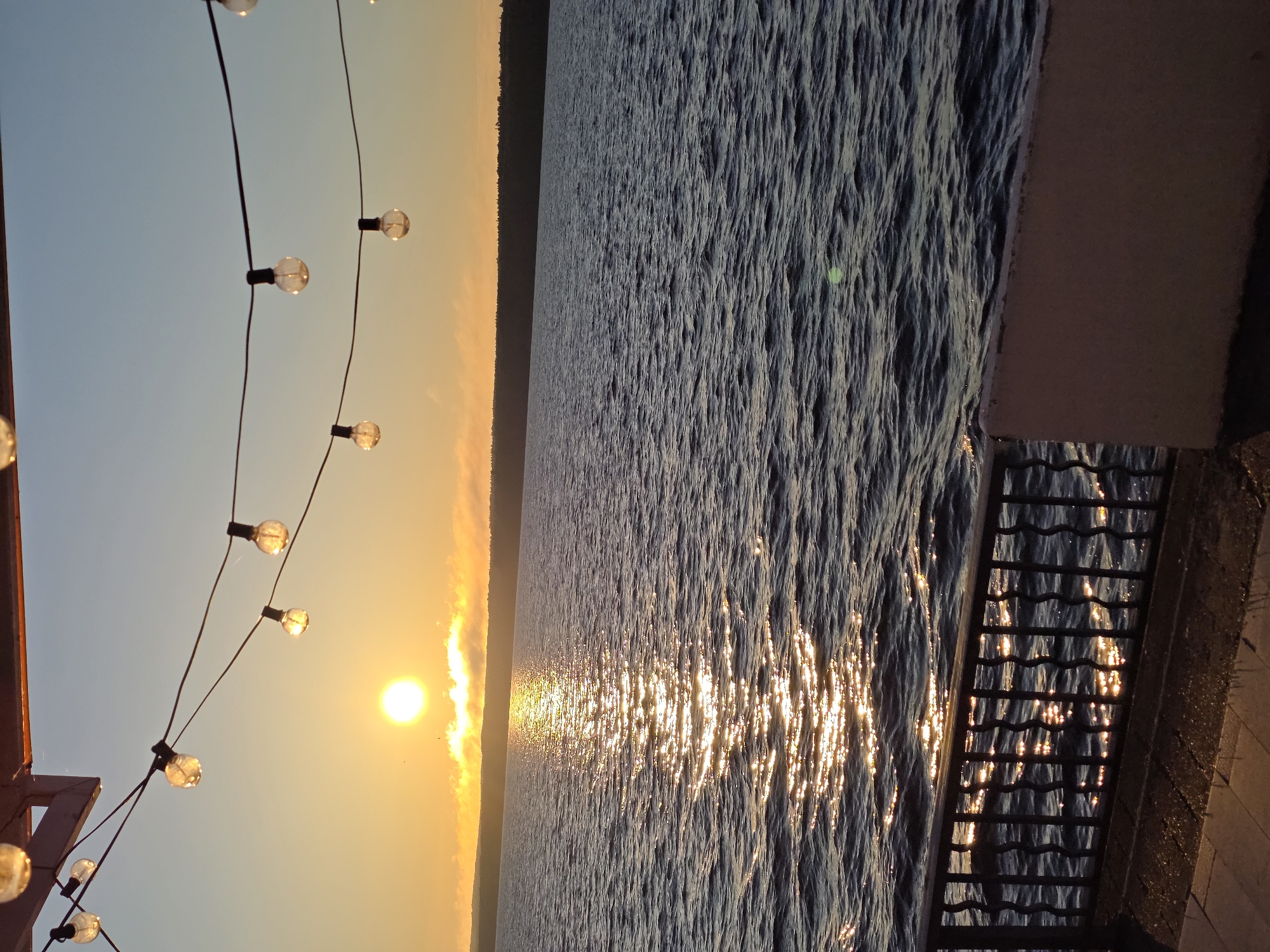 The image depicts a sunset over a body of water, likely a reservoir or lake, as seen from a paved area with a metal railing in the foreground. The sky transitions from a clear blue at the top to a warm orange and yellow hue near the horizon, illuminated by the setting sun. A string of decorative lights hangs across the top of the frame. The water is dark blue with ripples reflecting the sunlight, creating a shimmering path towards the shore. A treeline is visible on the distant horizon. The overall scene is peaceful and atmospheric. This scene could be located along the Kama River in Chaikovsky, Russia, as the city is situated on the banks of the river and is known for its reservoirs and industrial landscape which would be consistent with a sunset view over water. There are no people or animals visible in the image.