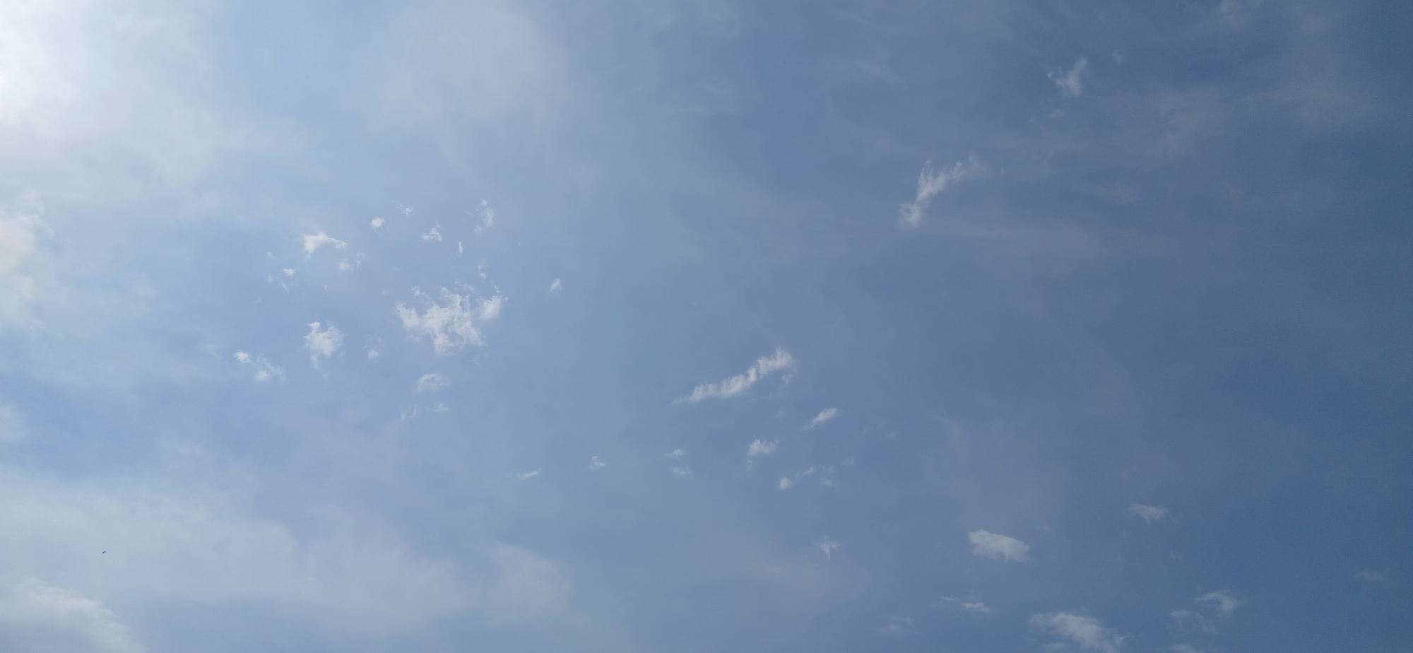 This is a video of a blue sky with scattered clouds. The sun is not directly visible, but its brightness casts a glow in the upper left portion of the frame, suggesting it is daytime. The clouds are wispy and thin, indicating fair weather. The overall scene is peaceful and serene, evoking a sense of calm. There are no people or objects visible in the frame, and no text is present. The location is unknown.