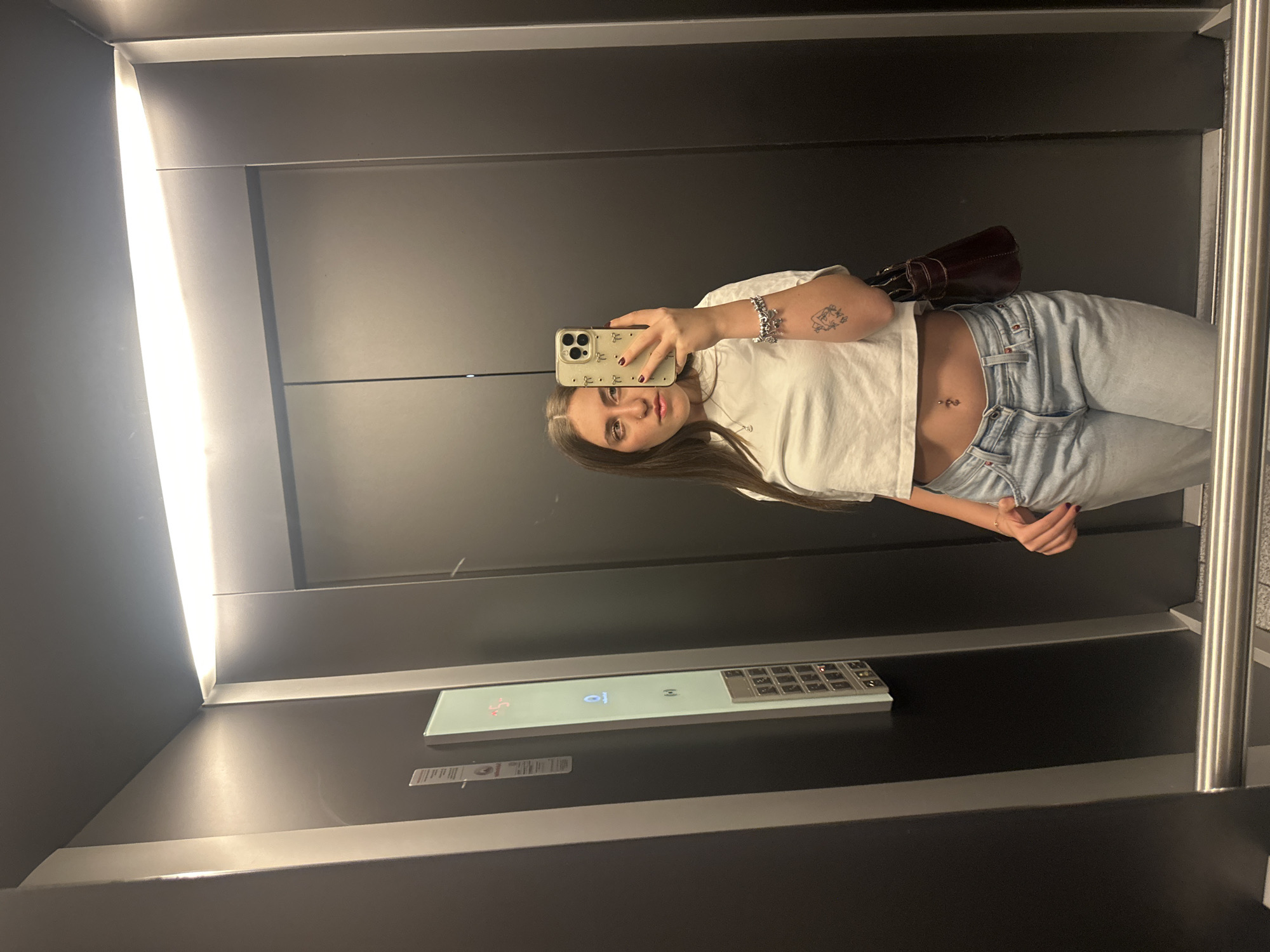 A young woman is taking a selfie in what appears to be an elevator. She is wearing a white t-shirt and light blue jeans. Her navel is pierced. She has a tattoo on her left arm. She is holding a smartphone with a light-colored case. A purse is visible over her left shoulder. The elevator has a control panel with buttons and a digital display. The walls of the elevator are dark gray, with bright lighting on one side. The location cannot be confirmed based on the image alone.