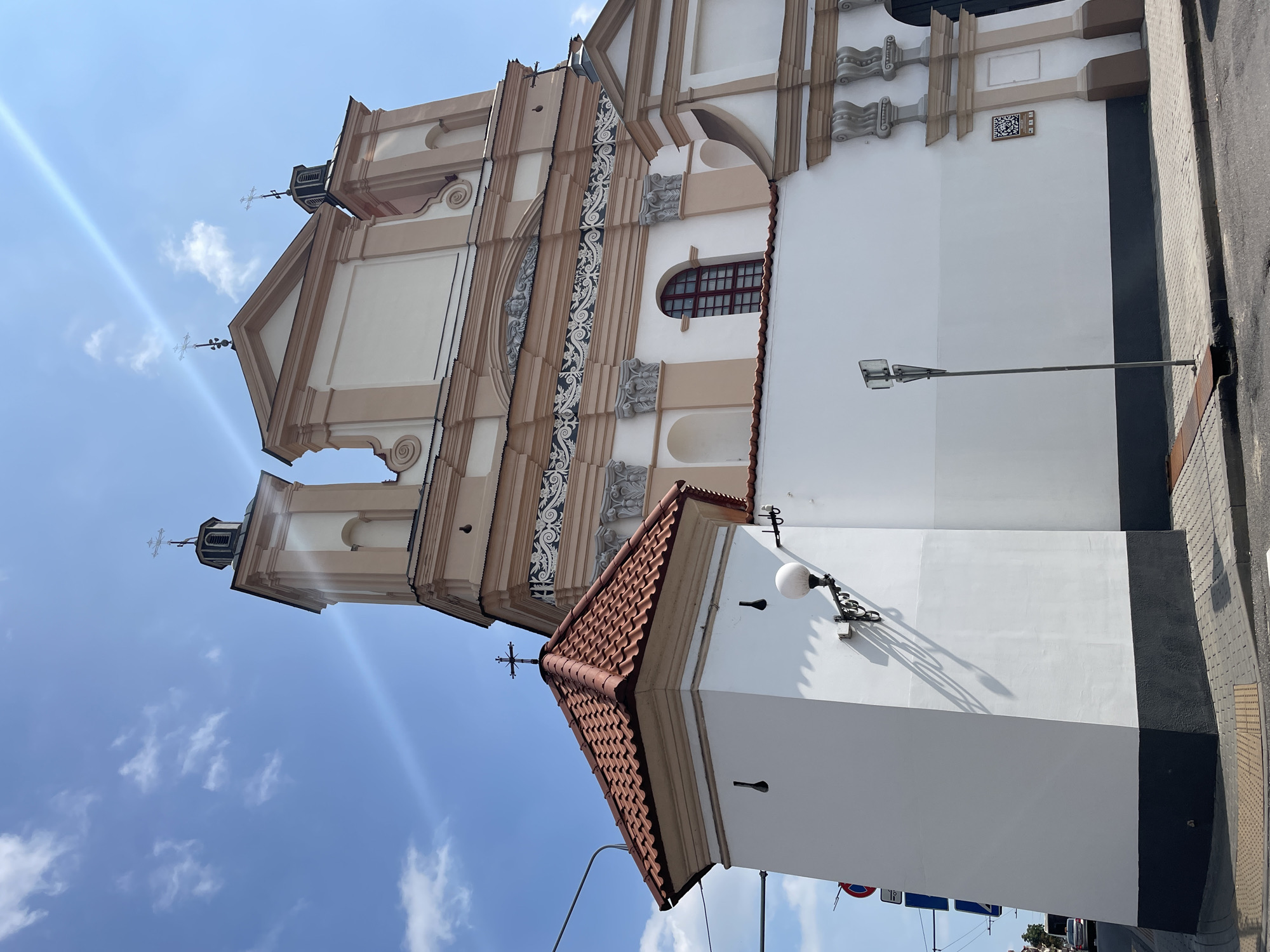 The image shows the Church of St. Francis Xavier in Hrodna, Belarus, on a sunny day with a blue sky and scattered clouds. The facade of the church is visible, with its distinctive Baroque architecture, including two towers topped with crosses. A smaller, white building with a red tiled roof and a cross on top is in the foreground to the left. The church is surrounded by a white wall, and a street with cobblestone pavement is in front of it. There are traffic signs and a lamppost visible on the sidewalk. A portion of a vehicle can be seen in the distance on the left side of the image. The overall scene depicts a religious building within an urban setting.