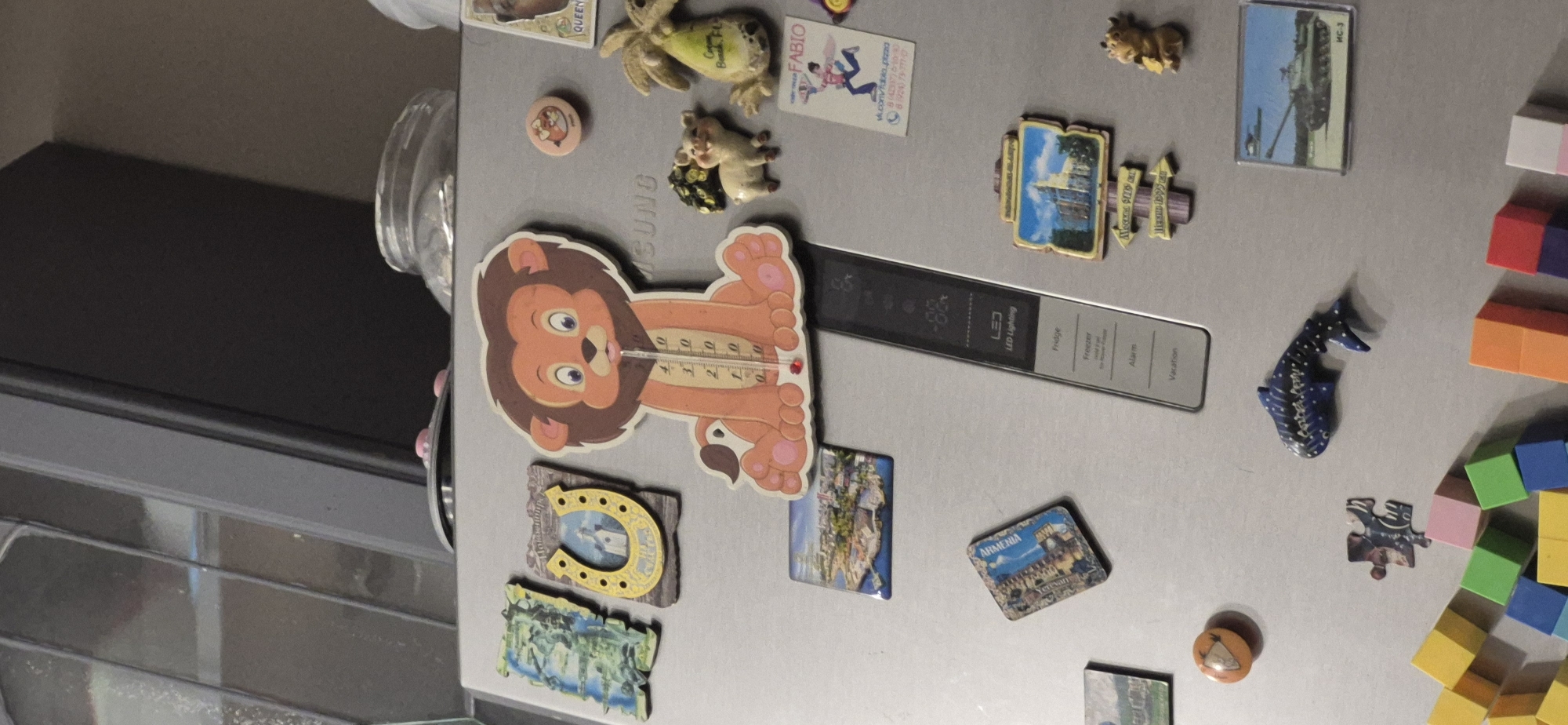 The image is a close-up, eye-level, indoor shot of a silver refrigerator door adorned with various magnets. The magnets include a cartoon lion with a thermometer, a horseshoe magnet with a church depicted inside, a small pig, a banana-shaped magnet, a small cow-like figurine, a magnet with a tank, a whale-shaped magnet, a jigsaw puzzle piece, and several colorful building block-shaped magnets scattered at the bottom. There are also several rectangular magnets depicting various landmarks and scenes, including a cityscape, a historical building with directional signs, and a postcard-style magnet featuring a person with text. A panel with "LED Lighting", "Fridge", "Freezer", "Alarm", and "Vacation" settings is visible on the right side of the refrigerator door. The background shows a reflection of a room, possibly a kitchen, with glass cabinets and a dark appliance. There are no people or animals present in the scene, and no actions are taking place. The location is not specified, and therefore is Unknown, Unknown.