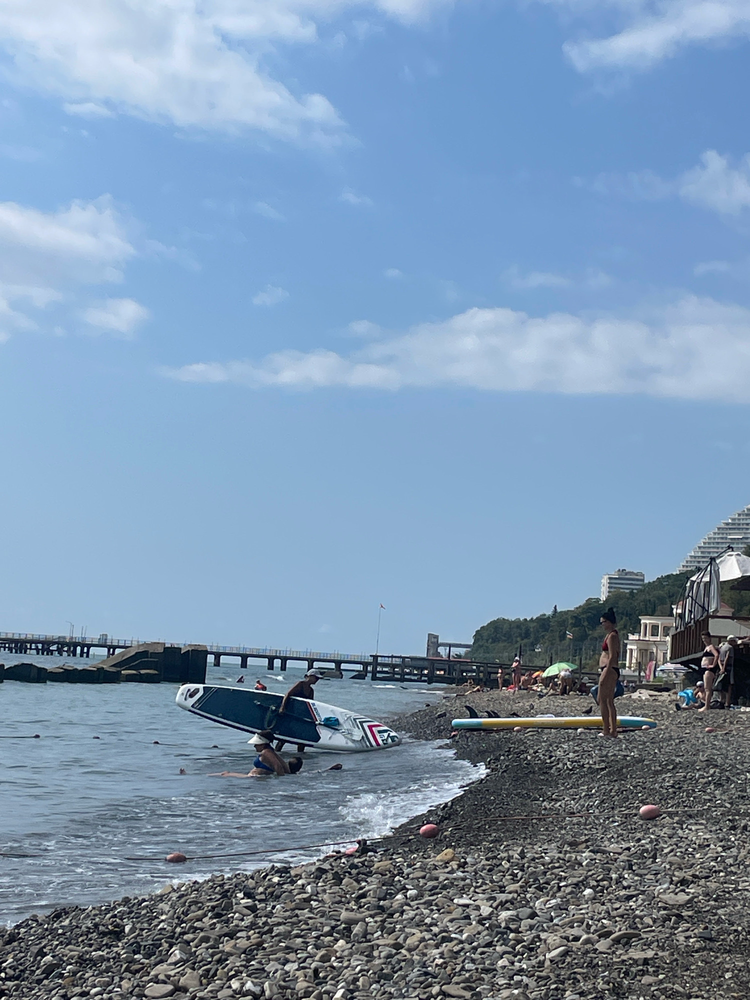 The image depicts a sunny day at a pebble beach in Sochi, Russia, with the Black Sea on the left and a built-up coastline on the right. In the foreground, two stand-up paddleboards are visible, one partially submerged in the water with two people in it, and another on the shore. Several other people are scattered on the beach, some sunbathing and others standing. In the background, a pier extends into the sea, and resort buildings are visible on the hillside. The sky is blue with scattered clouds.