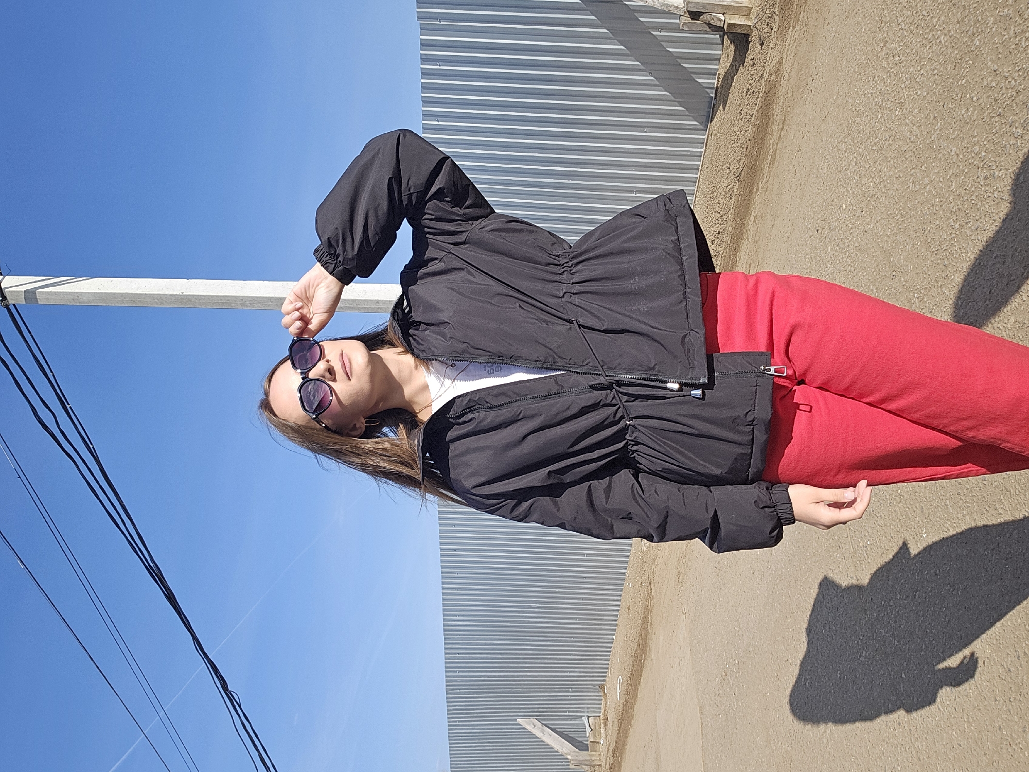 An outdoor, full-body shot depicts a woman standing on a dirt or gravel path under a clear blue sky. She is oriented slightly towards the left of the frame, looking up and towards the viewer. She wears a black, cinched-waist jacket over a white top and bright red pants. Dark sunglasses cover her eyes, and her right hand is raised to adjust them. Her brown hair frames her face.
Behind her, to the right, is a tall, corrugated metal fence, suggesting an industrial or utilitarian area. To her left, the ground is a light brown, dusty surface, and a long, distinct shadow of her body is cast, indicating bright sunlight. Several black power lines crisscross the upper left portion of the blue sky. A small wooden structure is visible near the base of the fence on the ground.
The image conveys a clear, sunny day, likely midday or early afternoon, given the sharp shadows and cloudless sky. The setting, located in Chaikovsky, Russia, appears to be an open outdoor space with functional elements like the fence and power lines. There is no visible text in the image.