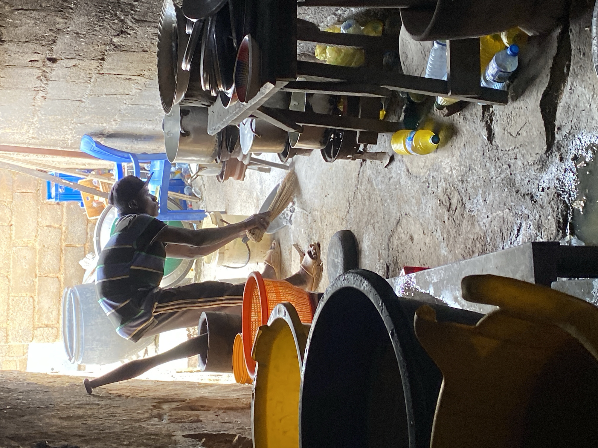 The image depicts a person sweeping a floor with a broom in a cluttered, indoor setting. The person, a young man wearing a striped t-shirt and a cap, is positioned in the lower-left quadrant of the frame, bent over and actively engaged in sweeping. His legs are visible, and he appears to be wearing sandals.
The scene is characterized by a variety of objects scattered around. There are large plastic containers and buckets, one of which is orange and appears to be a sieve or basket. Other containers are black and yellow. Behind the person, a blue chair is visible, and further back, a shelving unit holds numerous pots and pans, suggesting a kitchen or food preparation area. Several plastic bottles, some filled with clear liquid and others with yellow liquid, are also present on the shelves and the floor. The floor itself appears to be made of concrete or packed earth, and parts of it are wet, with visible puddles and some debris. The walls are constructed of rough, rough-hewn stones, characteristic of older buildings.
The lighting suggests it might be daytime, with natural light entering from an unseen source, casting some shadows. The overall atmosphere appears to be one of work and possibly a somewhat disorganized environment. The image's orientation is sideways, with the action unfolding from right to left. There is no visible text in the image. The location context of Jos, Nigeria, hints at a potentially hot climate, though this cannot be definitively determined from the image alone.