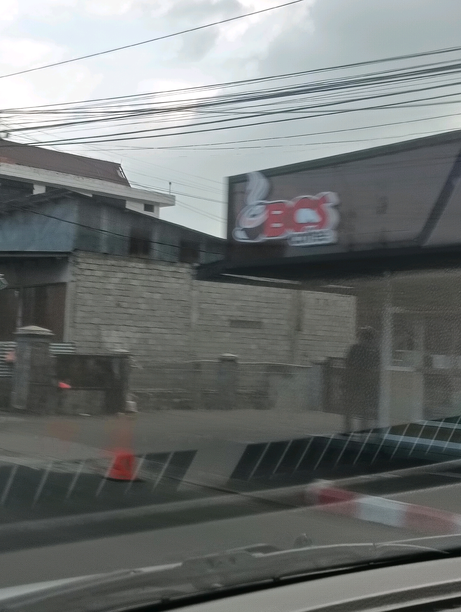 The image captures a scene from a street in Tomohon, Indonesia. In the foreground, a blurry view through a vehicle's windshield shows a road surface with diagonal metal grates and a red and white barrier. Further ahead, an orange traffic cone is visible. In the mid-ground, a building with a grey brick facade is situated behind a fence. Above this building, a sign with red lettering and a cartoon character is affixed to a dark structure. The background features overcast skies and a network of overhead power lines. A shadowy figure is discernible through a glass panel on the right side of the mid-ground building.