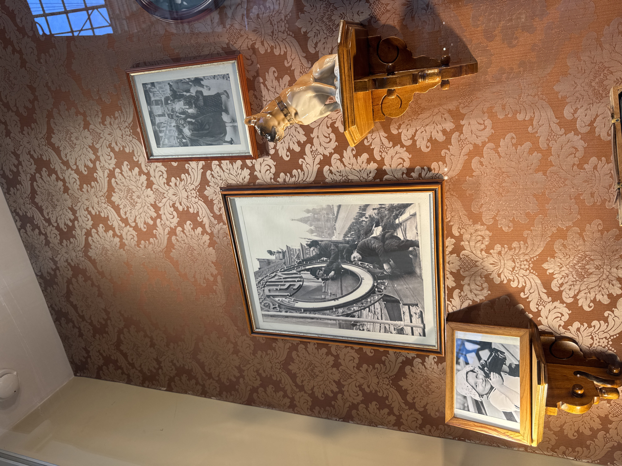 The image displays a wall adorned with a patterned, reddish-brown wallpaper. Several framed photographs and a decorative statuette are mounted on the wall.
In the center of the frame, a large, black and white photograph depicts a group of men working on a large, circular clock face. One man is positioned at the top of the clock, appearing to adjust something, while another man kneels below him. The background of this photograph shows buildings, including what appears to be Saint Basil's Cathedral in Red Square, suggesting the location is Moscow, Russia.
To the upper right of this central photograph, a smaller, framed black and white image shows a crowd of people walking along a street.
Below the large clock photograph and to the left, a smaller framed photograph shows a young child wearing a hat and holding a camera. This photograph is mounted on a wooden shelf.
To the right of the large clock photograph, a statuette of a dog, possibly a bulldog, sits on a wooden shelf. The dog is light brown and white, wearing a dark collar. The wooden shelf it rests on is ornately carved.
The overall impression is of a wall decorated with historical images and a decorative object, possibly in a home or museum setting in Moscow. The lighting suggests an indoor environment.