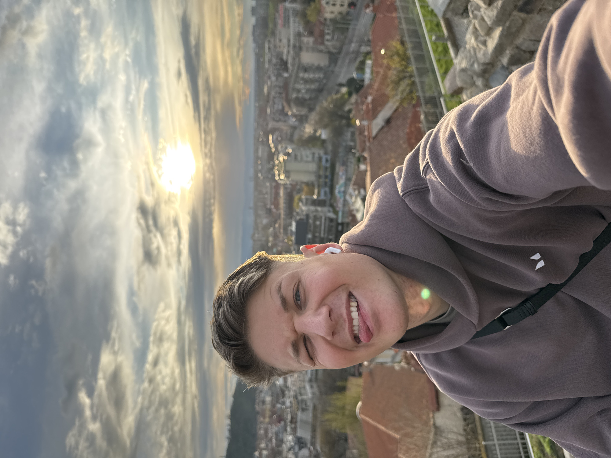A close-up selfie of a young man with short, light brown hair is taken against the backdrop of Plovdiv, Bulgaria. He is wearing a brown hooded sweatshirt and has an in-ear wireless earbud in his right ear. The man is winking with his left eye and sticking out his tongue, suggesting a playful or jovial mood. The background reveals an aerial view of the city, with the setting sun diffused through the clouds, casting a warm glow on the landscape. Buildings and streets are visible, indicating an urban environment. The man appears to be positioned at an elevated location within the city, possibly a viewpoint or high building, as suggested by the perspective of the city below. The image orientation is somewhat off-kilter as the man is tilted to the right.