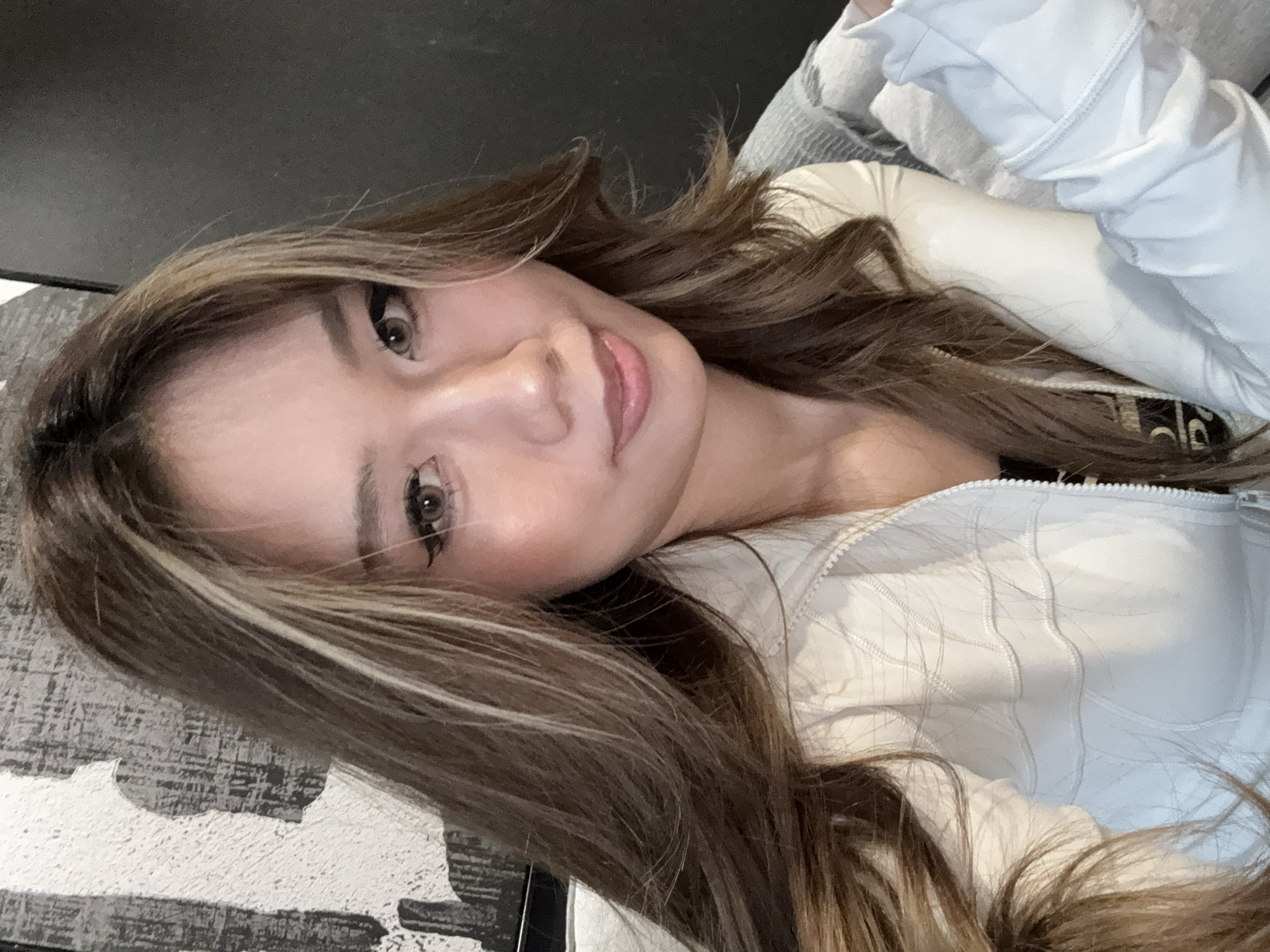 A woman with long, wavy brown hair with blonde highlights is looking at the camera. She is wearing a white jacket. The background is a mix of a patterned surface and a dark, undefined area. There is no information to indicate the location of the image is in the city of Liangjiang, China.