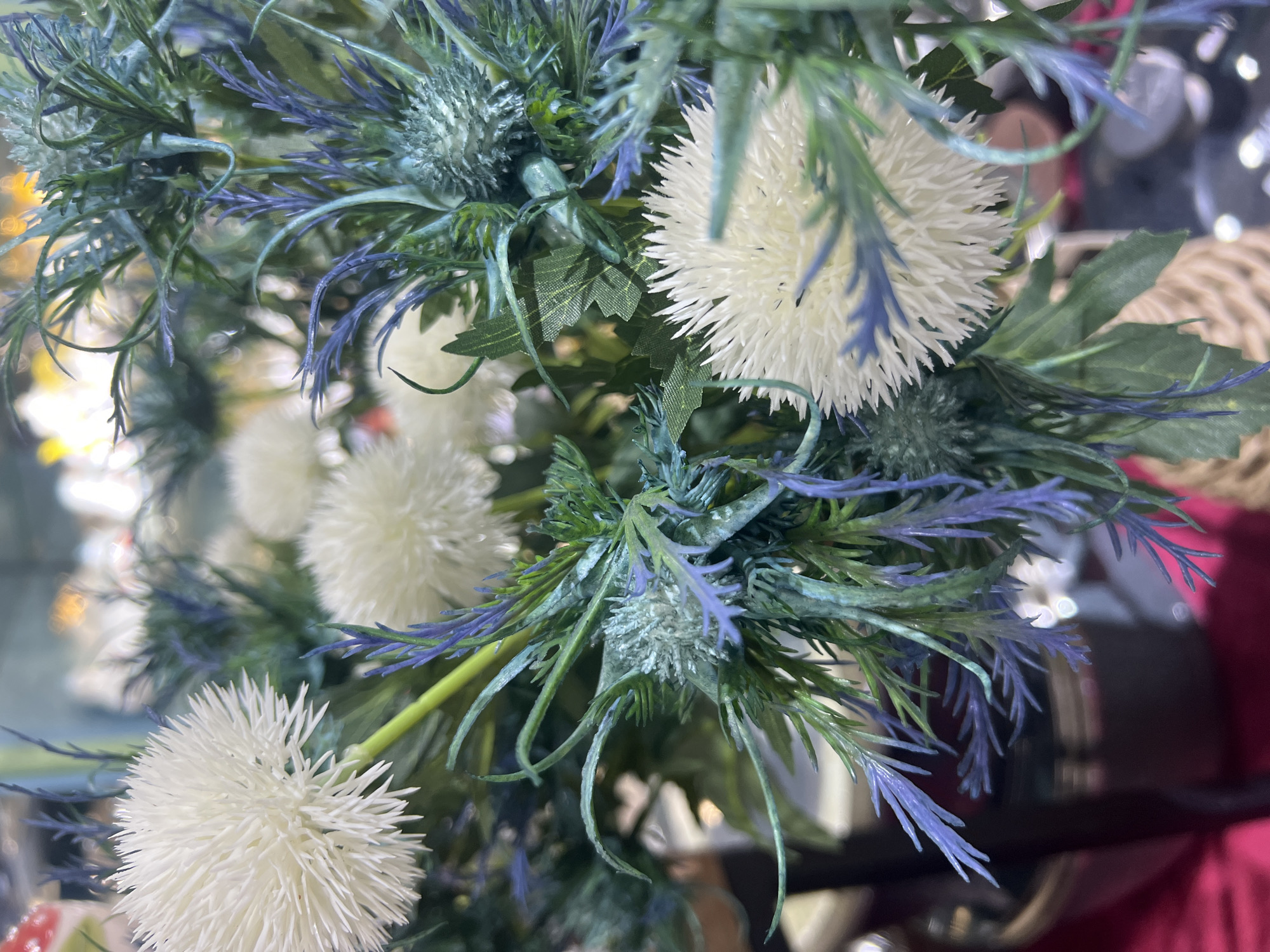 The image displays a close-up view of a floral arrangement, likely artificial, featuring white, spherical blossoms resembling dandelions or thistles, interspersed with spiky, blue-green foliage. The background is blurred, suggesting a shallow depth of field, with hints of reflective surfaces and other out-of-focus objects, possibly part of a retail or decorative setting. There are no people or identifiable landmarks present. The location in Eshanguzar, Uzbekistan, is not visually represented in the provided image.