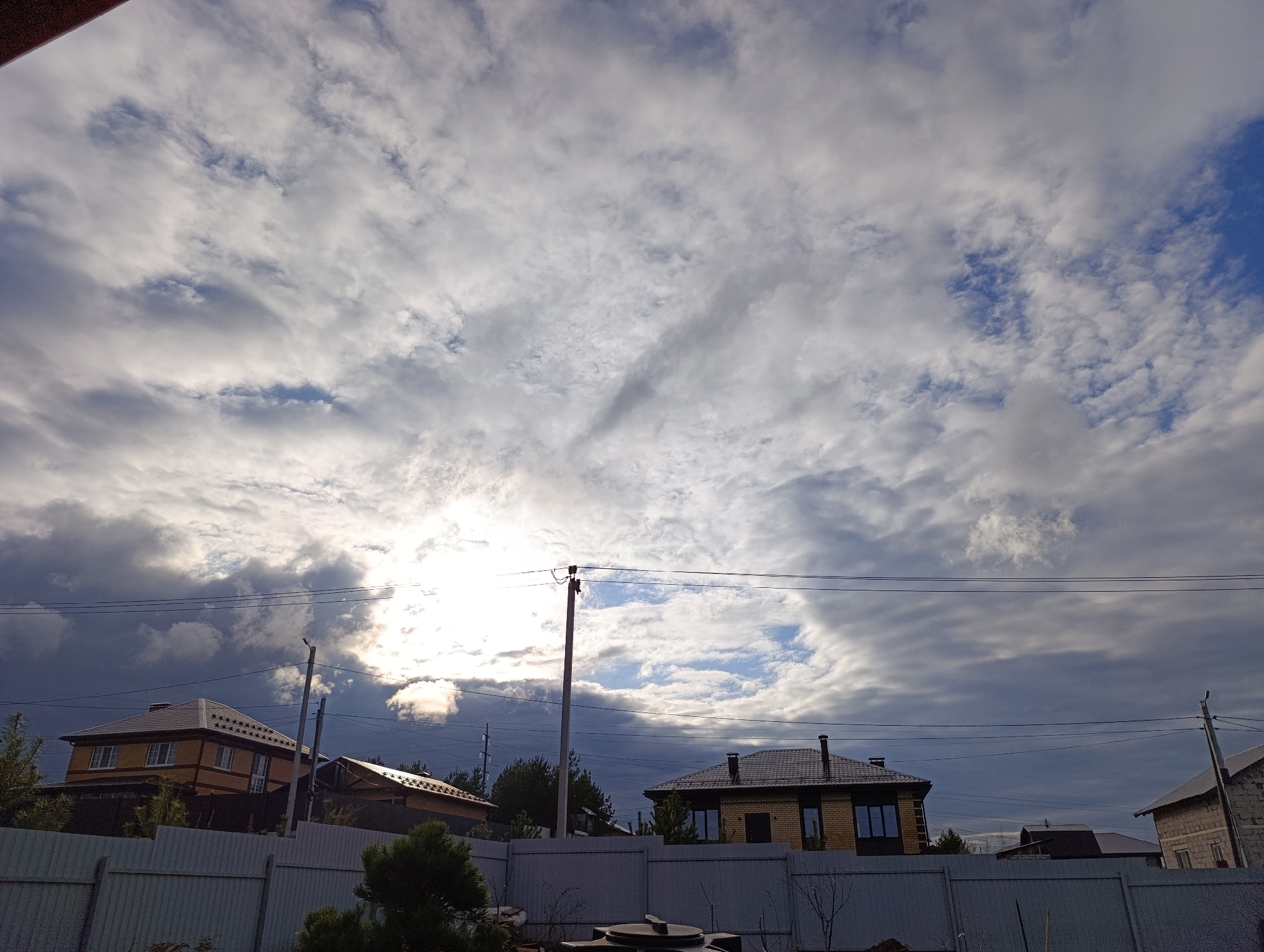The image shows a cloudy sky over a residential area in Chaikovsky, Russia. Several houses with varied architectural styles are visible behind a grey fence. Power lines traverse the scene in the mid-ground. The sky is mostly covered with grey and white clouds, with a patch of blue sky breaking through. The sun is obscured by the clouds, creating a diffuse, bright area. In the foreground is a white fence. Some plants and other objects can be seen behind the fence.