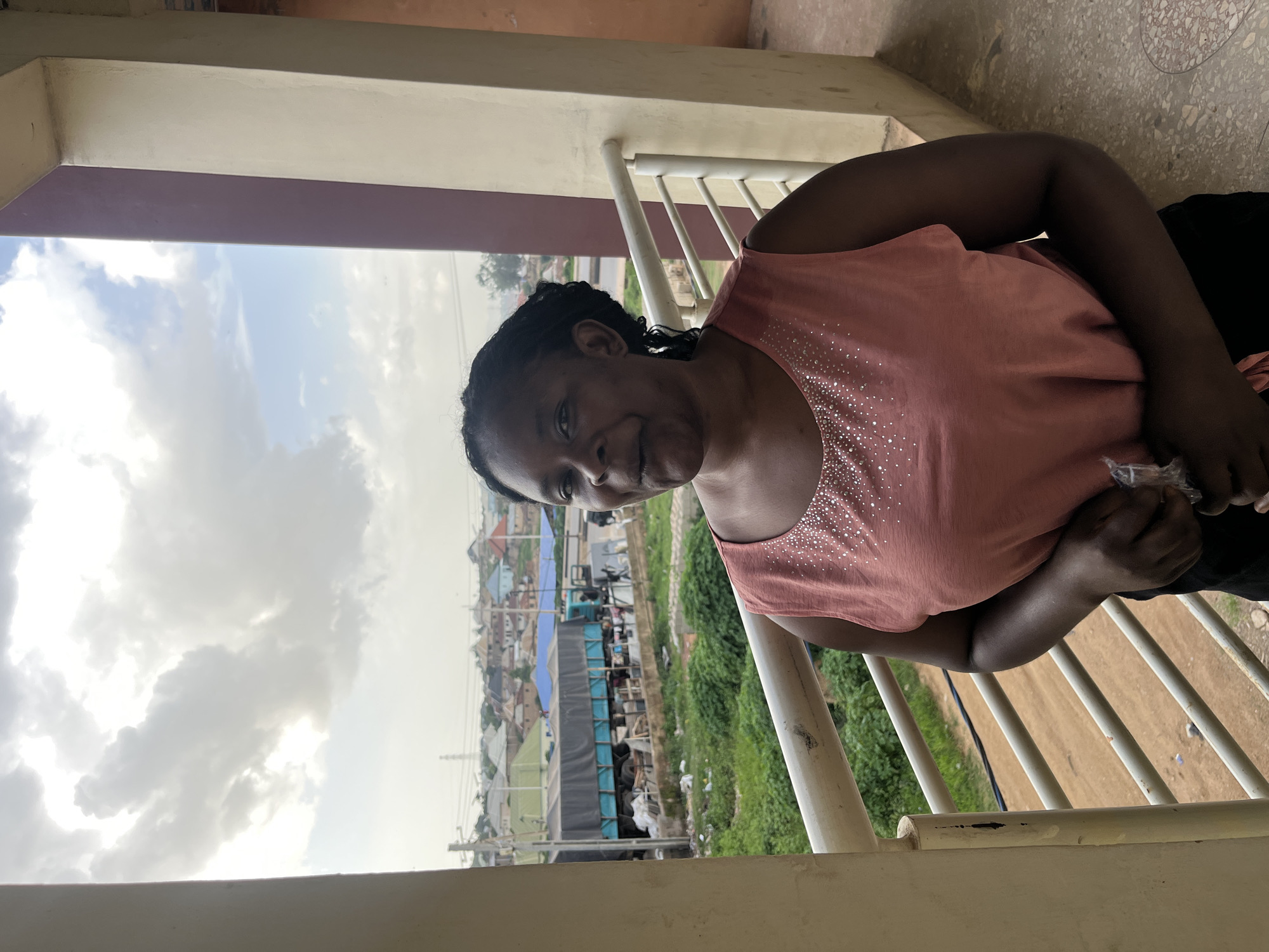 A medium shot photograph captures a Black woman standing on a balcony in Gura Top, Nigeria, under a cloudy sky. She is positioned on the right side of the frame, facing forward with her head turned slightly to her left, looking towards the camera with a neutral expression. Her braided dark hair is pulled back, and she is wearing a sleeveless, coral-pink top adorned with small, clear rhinestones around the neckline. Her arms are crossed in front of her, and she holds a transparent plastic bag in her hands.
The balcony features a white railing with vertical supports. Beyond the railing, the scene unfolds to reveal a cityscape. Green foliage and what appears to be an unpaved road are visible below, with scattered debris. Further in the distance, a blue-covered truck is parked, and a cluster of multi-colored buildings with various roof shapes and colors make up the urban landscape. The sky is dominated by large, white and gray clouds, with patches of blue sky peeking through. The overall lighting suggests it is daytime. The right edge of the frame shows a textured wall, possibly part of the building the balcony is attached to.