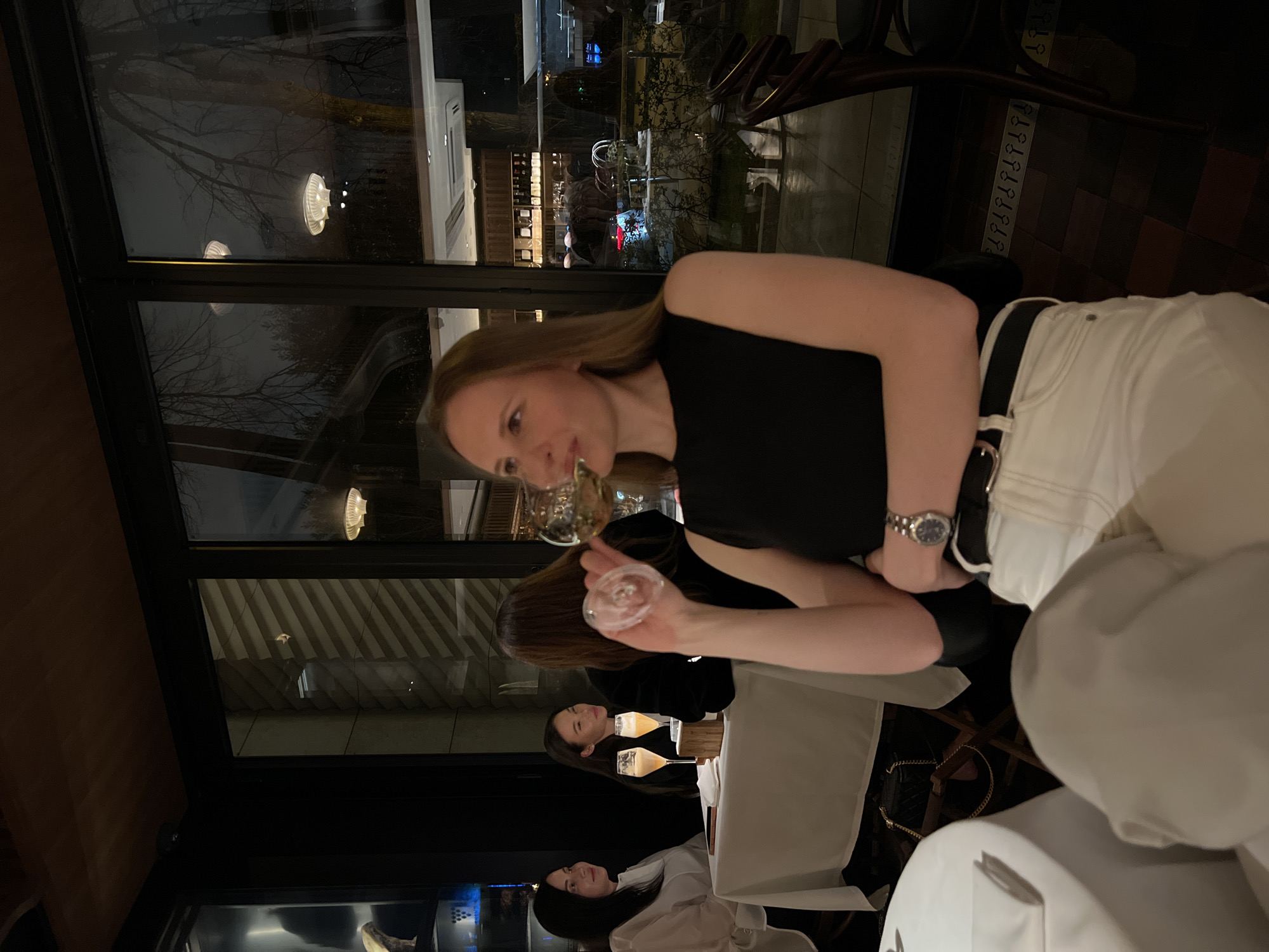 The image shows a group of people at a restaurant in an unknown city and location. The restaurant has large windows with a view of the outside, including trees and buildings. One woman in the foreground, wearing a black top and white pants with a black belt, holds a glass. Another woman is visible in the background, also dressed in black. A third woman, wearing a white shirt, is visible at the far end of the table. The table is covered with a white tablecloth and has multiple glasses on it. There are chairs around the table, and a small black purse hangs from one of them.