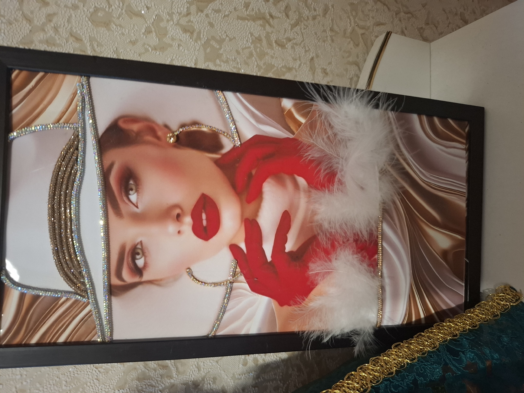 This is a framed artwork featuring a close-up of a woman's face. The woman has striking makeup, with bold red lips and dramatic eye makeup. She is wearing a white hat adorned with rhinestones and gold trim. Her hands are also covered in red gloves, and she is surrounded by white feathers. The background of the artwork is abstract, with swirling patterns of brown and gold. The artwork is displayed in a dark frame and is positioned against a textured, light-colored wall. A portion of a decorative gold trim is visible in the foreground. The overall impression is one of glamour and artistry. The specific location of Jitiqara, Kazakhstan, is noted, but there are no visual cues within the image to confirm or suggest this location. No text is visible in the image.