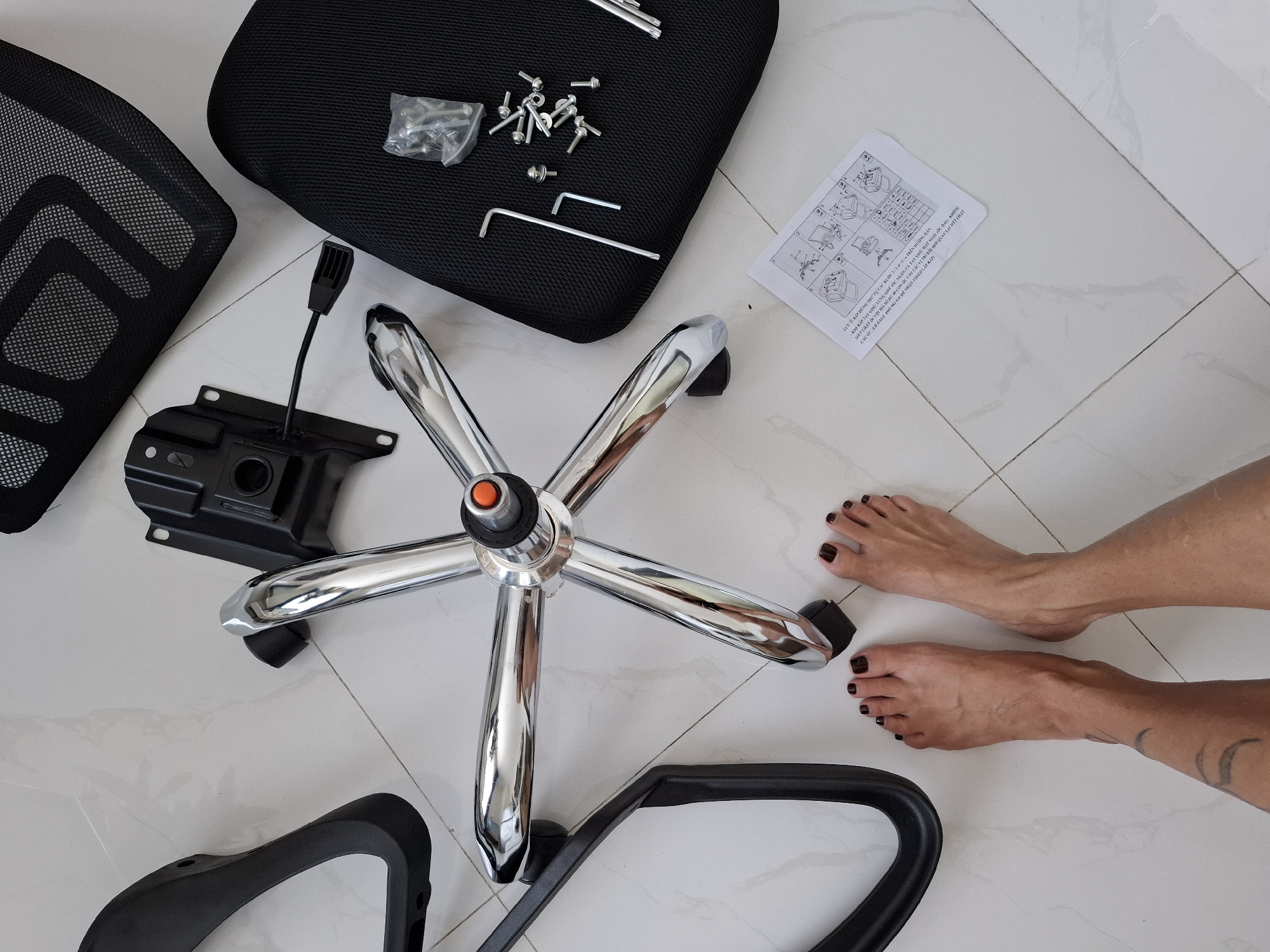This is an overhead, full-shot, eye-level, indoor photograph of office chair parts scattered on a light-colored tiled floor. The parts include a chrome five-pronged base with wheels, a black seat cushion, a black mesh backrest, a black armrest, and the chair's mechanism. Several screws and two Allen wrenches are also visible, along with an instruction manual for assembling the chair. A pair of feet with dark red nail polish are positioned in the lower left quadrant of the image, suggesting a person is present but not fully visible. The lighting is bright and appears to be natural daylight. The scene is consistent with a home or office environment, not specific to Nha Trang Ward, Vietnam.