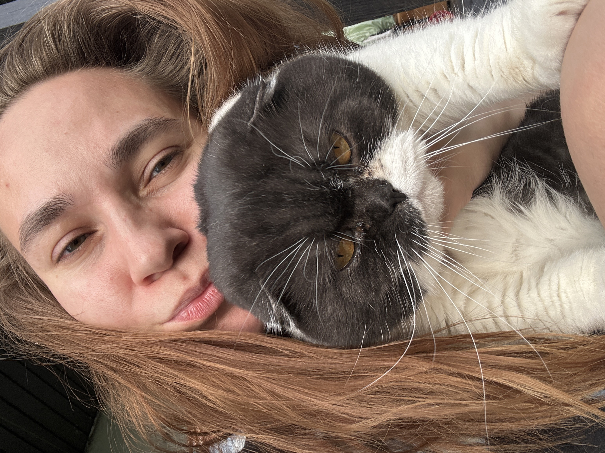 A woman with light brown hair is lying down, with her head resting on the left side of the frame. She is embracing a black and white Scottish Fold cat. The cat is mostly black on top of its head and white on the lower part of its face and body. The woman has light skin and pursed lips.