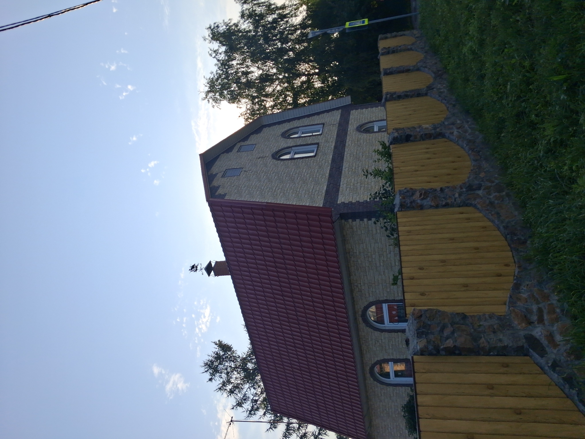 This is a medium-distance, eye-level, outdoor, daytime, full-shot photograph taken in Novosibirsk, Russia. The image captures a two-story house with a red, tiled, gambrel roof. The house's exterior walls are made of light-colored, rectangular stone veneer, with a darker stone band running horizontally below the upper windows. The house features arched windows on both levels and a small rectangular window on the upper left side. A brick chimney with a weather vane shaped like a ship stands on the roof. The foreground is dominated by a wooden fence with stone pillars, built on a grassy slope. In the background, mature trees with dark green foliage frame the right side of the house, and a traffic sign is visible on the right. The sky is a clear, light blue with scattered, wispy clouds. There are no people or animals visible in the image.