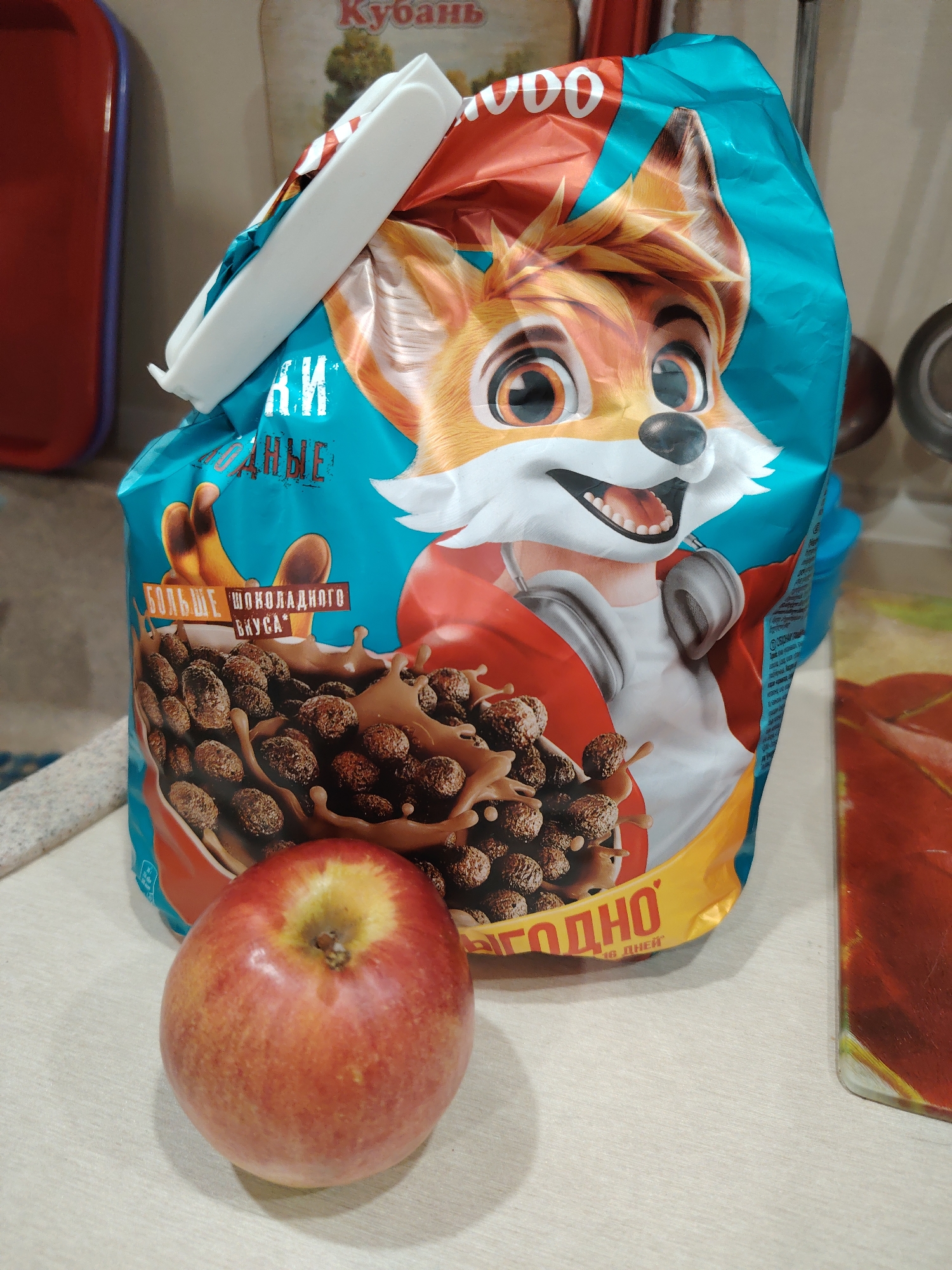 A forensic image depicts a domestic indoor setting, likely a kitchen or pantry, with a large bag of cereal and a single apple positioned on a light-colored countertop. The central object is an upright, blue and red cereal bag, partially crinkled, featuring a cartoon fox character wearing headphones. The bag's opening is folded down and secured by a white plastic bag clip. Russian text is visible on the bag, including "БОЛЬШЕ ШОКОЛАДНОГО ВКУСА" (More Chocolate Flavor) and "ВЫГОДНО" (Profitable/Beneficial) along with a graphic of chocolate cereal balls in a bowl with milk. Directly in front of the cereal bag, slightly to the left, rests one whole red apple, exhibiting red and yellow color variations. Both items are situated on a light beige, textured countertop. In the blurred background, a red plastic container is partially visible on the far left, and a red and yellow object, possibly a placemat, is present on the right. Further back, a light brown object with the red Cyrillic text "Кубань" is partially discernible. No individuals are present. The location is Unknown, Unknown.