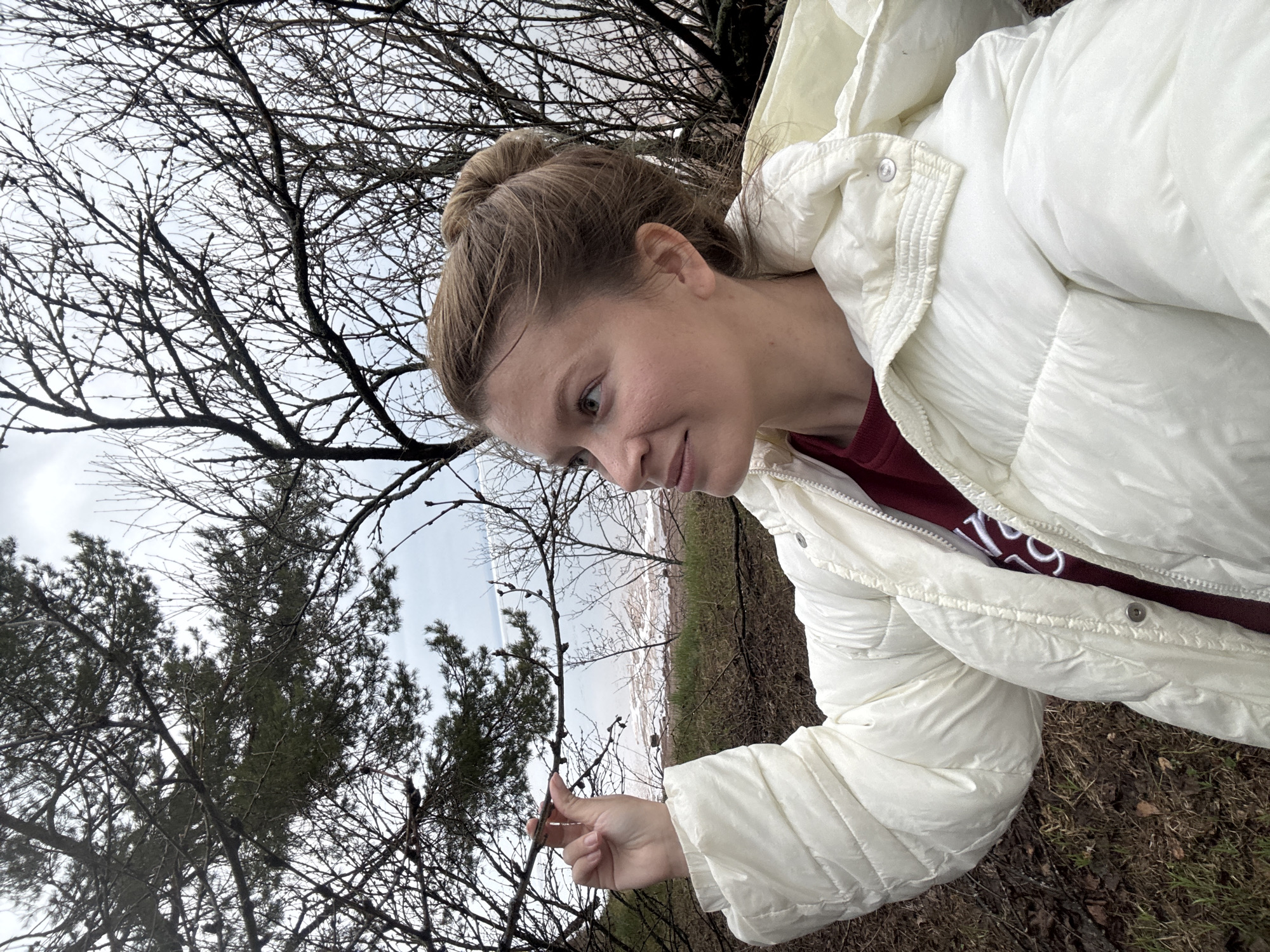 A person is outside, likely in a park or natural area. The person has their hair pulled back in a bun and is wearing a white puffer jacket over a maroon shirt. They are holding a small twig with buds on it in their hand. The background consists of bare trees and some greenery, with a glimpse of a light-colored area possibly water or a beach in the distance. The ground appears to be covered in dry leaves and grass. The photo seems to have been taken at a low angle or slightly tilted. The location is not discernible from the image itself.