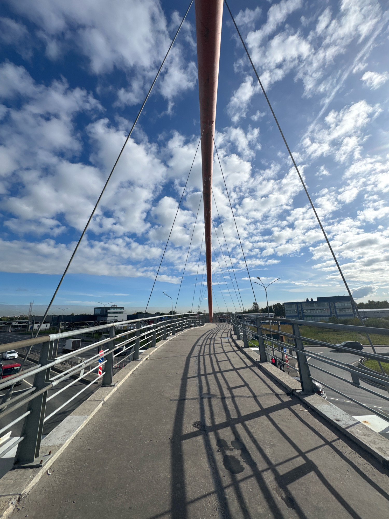 A low-angle, wide-shot perspective captures a pedestrian bridge in Saint Petersburg, Russia, on a bright, cloudy day. The curved, concrete walkway of the bridge is in the foreground, leading the viewer's eye towards the horizon. A metallic railing lines the sides of the walkway, casting distinct, geometric shadows across the path due to the angled sunlight.
Dominating the upper portion of the frame is a vibrant blue sky, dotted with numerous white, cumulus clouds. Sunlight streams from the right, creating a bright halo effect and illuminating the scene. A large, red, curved pylon, a key structural element of the bridge, rises prominently from the left side of the image, its surface slightly textured. Several suspension cables extend diagonally from this pylon towards the unseen anchor points, crisscrossing the sky.
In the middle ground, the bridge overlooks a multi-lane road with ongoing traffic, including cars and trucks. Buildings, including a modern office building with several stories and a telecommunications tower, are visible in the distance, interspersed with trees and greenery. Streetlights are positioned along the road. The overall environment suggests an urban setting, likely a suburban area or a transport hub on the outskirts of the city. No people are visible in the image.
