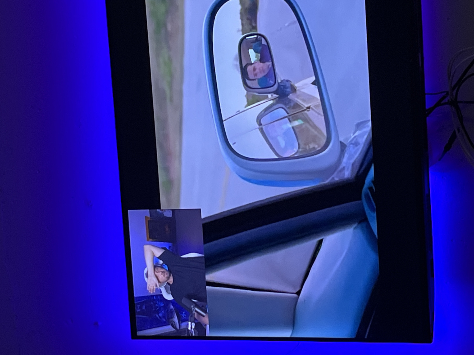 The image displays a split-screen view, likely from a video feed. The left side shows a man in a black t-shirt and a white cap, seated in a gaming chair, with his hand covering his forehead. He appears to be in a room with computer equipment and a framed picture in the background. The right side of the image features a view through a car's side mirror, reflecting another man. This man, wearing a purple shirt, is seated in the driver's seat of a car and is looking forward. The background outside the car shows a paved road and distant trees. The television displaying this content is framed by blue LED lighting, suggesting it is in a dimly lit room. There is no discernible connection to the city of Tomsk, Russia.
