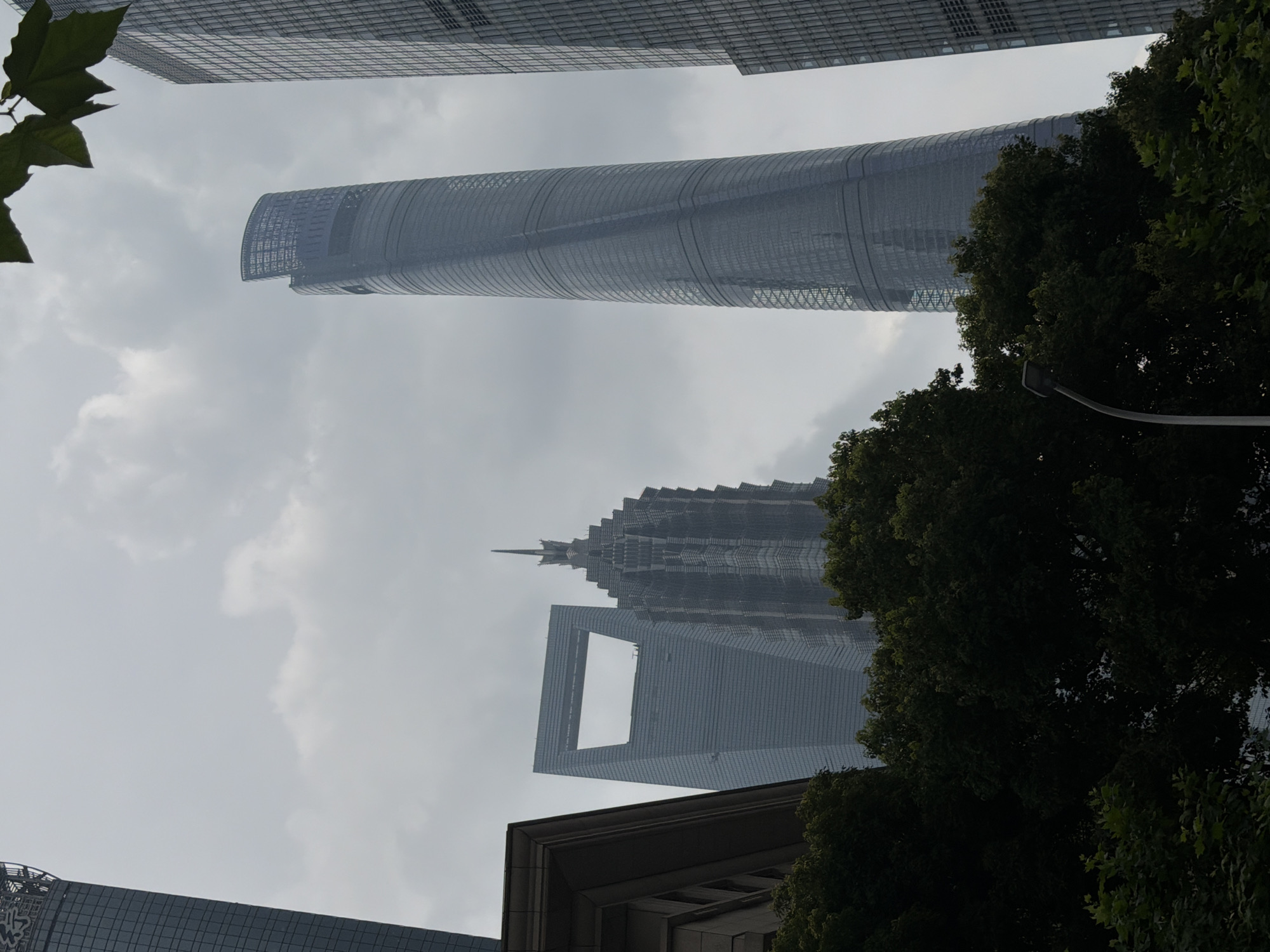 This is a low-angle shot of several modern skyscrapers in Pudong, China, under a cloudy sky. In the foreground, lush green trees obscure the lower portions of the buildings. On the left, a dark building with stone accents is partially visible. To its right, a glass skyscraper with a distinctive rectangular opening at the top rises. Further to the right is a complex, spired skyscraper. The most prominent building in the center-right is a tall, curved, glass skyscraper, appearing to have a spiraling pattern on its surface. To the far right, another tall, rectangular glass skyscraper is visible. A streetlamp on a curved pole emerges from behind the trees on the right. No people are visible in the image.