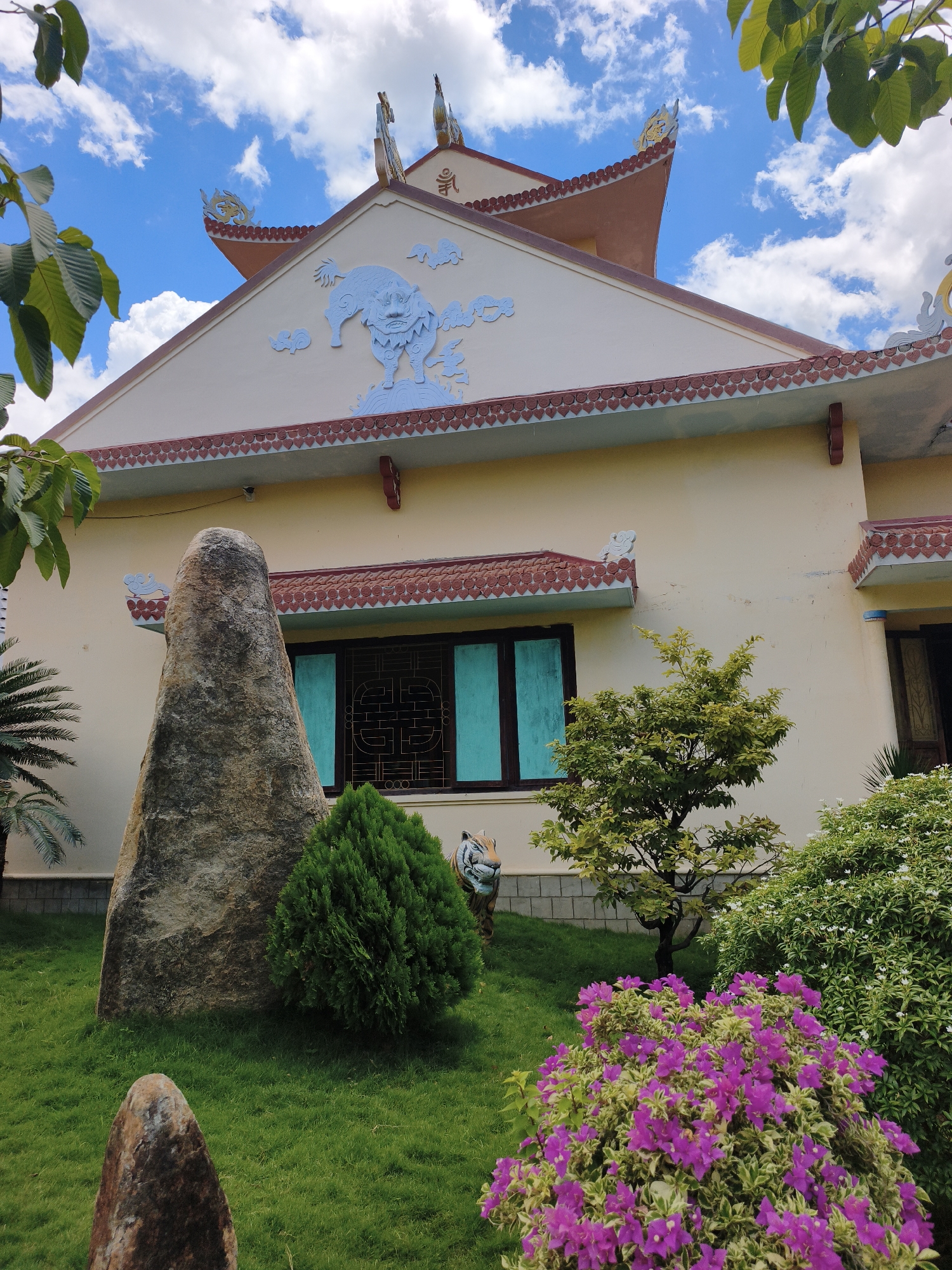 The image depicts a Buddhist temple or pagoda in Cam Ranh, Vietnam, characterized by its distinct architecture and lush surrounding garden. The temple features a pale yellow facade with a reddish-brown tiled roof, adorned with decorative patterns. A prominent element of the facade is a large blue sculpted dragon or mythical beast, and there are also bird-like figures on the roof's apex. In the foreground, a large, weathered, conical stone stands on a vibrant green lawn. Adjacent to the stone is a manicured green evergreen bush. Further to the right, a small, leafy tree with lighter green foliage stands. In front of this tree, a cluster of bright pink bougainvillea flowers adds a splash of color. Behind the bougainvillea and to the right of the evergreen bush, a dense green shrub with small white flowers is visible. A portion of a tiger statue is partially obscured behind the evergreen bush. The sky above is a bright blue, filled with fluffy white clouds. The overall scene suggests a serene and well-maintained religious or spiritual site within a tropical environment.