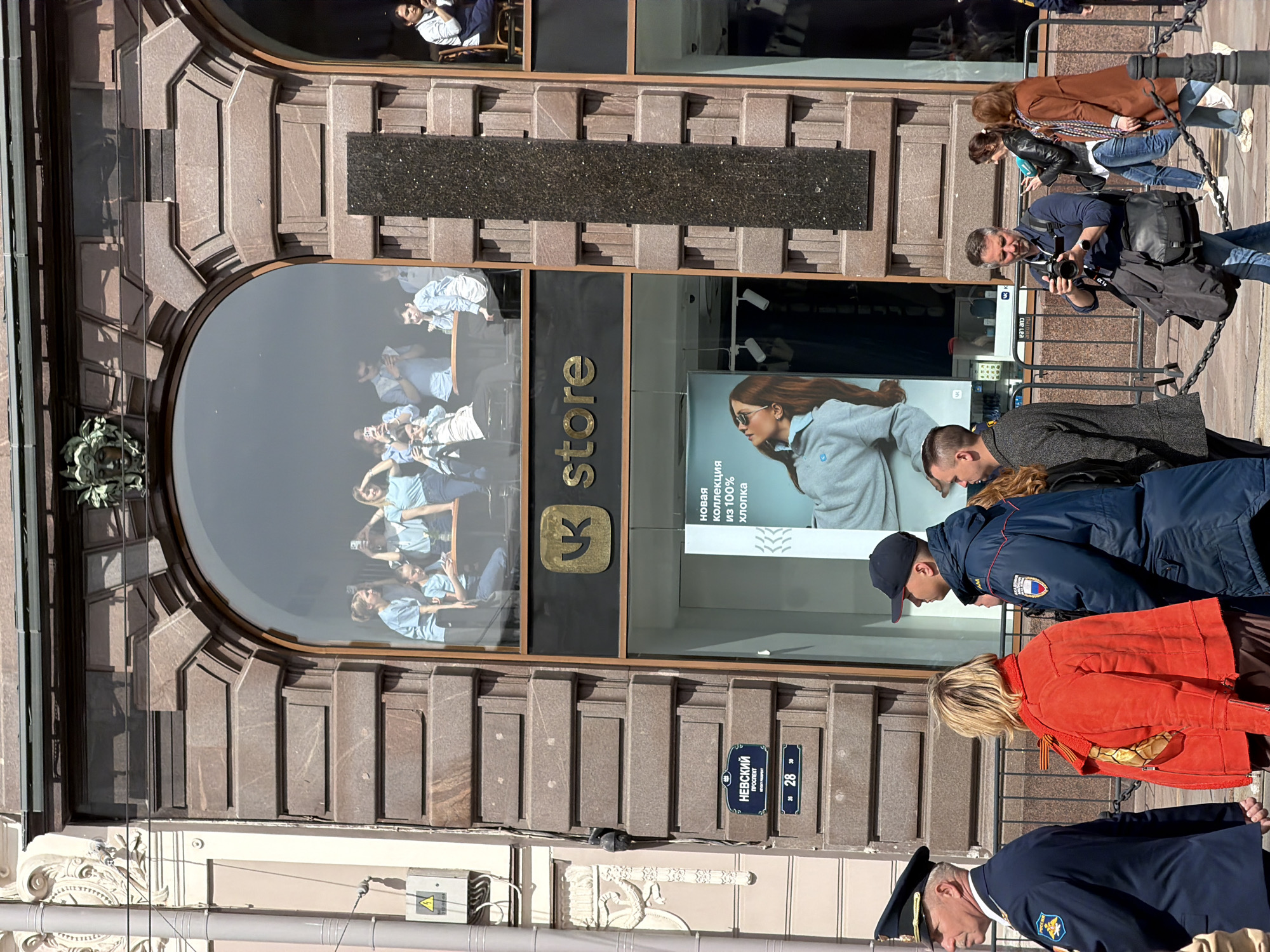 Here is a detailed description of the image:
**General Description:**
The image depicts a street scene in Saint Petersburg, Russia, focused on the exterior of a store. Several people are walking by the store, and a couple of uniformed officers are in the foreground.
**Location:**
The image is set on Nevsky Prospekt in Saint Petersburg, Russia. This is indicated by a street sign that reads "Nevsky Prospekt, 28, 30."
**Subjects and People:**
- Several pedestrians are walking along the sidewalk, including a man taking pictures with a camera, and people wearing jackets and coats.
- Two uniformed police or security officers are walking in the foreground.
**Objects:**
- The store's facade, including a large, arched window. Inside the store is the logo and word "store" of the Russian social media platform, VK (formerly VKontakte). A promotional poster is also visible in the store window.
- Street signs indicating the location on Nevsky Prospekt.
- Chain barriers along the sidewalk.
**Actions and Interactions:**
- People are walking and observing the store.
- The man is taking pictures of the store.
- The uniformed officers are walking along the sidewalk.
**Environment:**
- The architecture of the building is ornate, featuring detailed stonework.
- The weather appears to be sunny.