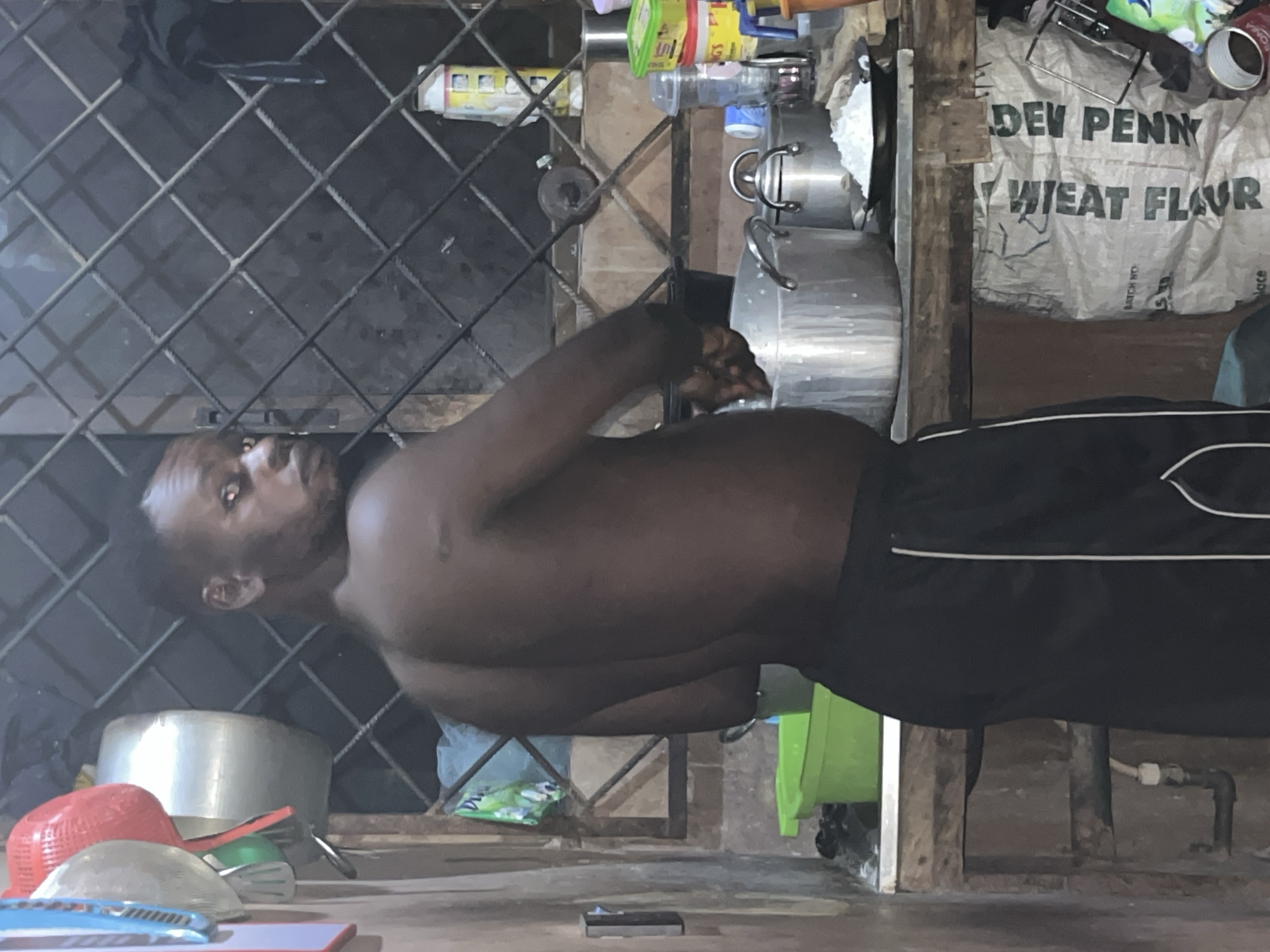 A shirtless Black man wearing black shorts with white piping stands in a kitchen, facing away from the camera and looking back over his left shoulder. He is positioned in front of a metal counter with several cooking pots and a plate of white rice. To his left, a green plastic basin and other kitchen items are visible. Behind him, a window with a metal grate is illuminated by an unseen light source. The kitchen appears to be in a state of disarray, with various containers and bags on shelves and the floor. A bag labeled "WHEAT FLOUR" is prominent on a lower shelf. The overall environment suggests a simple and functional, albeit cluttered, living space in a location identified as Unknown, Unknown.