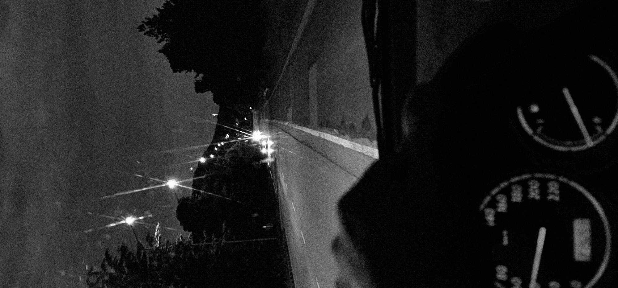 This is a grainy, black and white photograph depicting a nighttime scene, seemingly taken from inside a vehicle. The image is oriented vertically, with the left side showing the exterior and the right side showing the vehicle's interior.
On the left, a dark, textural sky occupies the upper portion, giving way to the stark silhouettes of tall trees and dense vegetation below. Numerous bright light sources with distinct starburst effects are scattered across this area, suggesting streetlights or distant vehicle lights. A narrow road or pathway runs vertically up the center of the left side of the frame, disappearing into the darkness at the top.
The right side of the image is dominated by the shadowed interior of a vehicle. In the bottom-right corner, two circular gauges from a dashboard are partially visible. The lower gauge, likely a speedometer, has a needle pointing to a low speed, with blurry numerical markings such as "100", "200", and "300" faintly discernible. The upper gauge is less clear.
The heavy grain and monochromatic palette contribute to a raw, perhaps vintage, aesthetic, conveying a sense of movement or travel during the night.