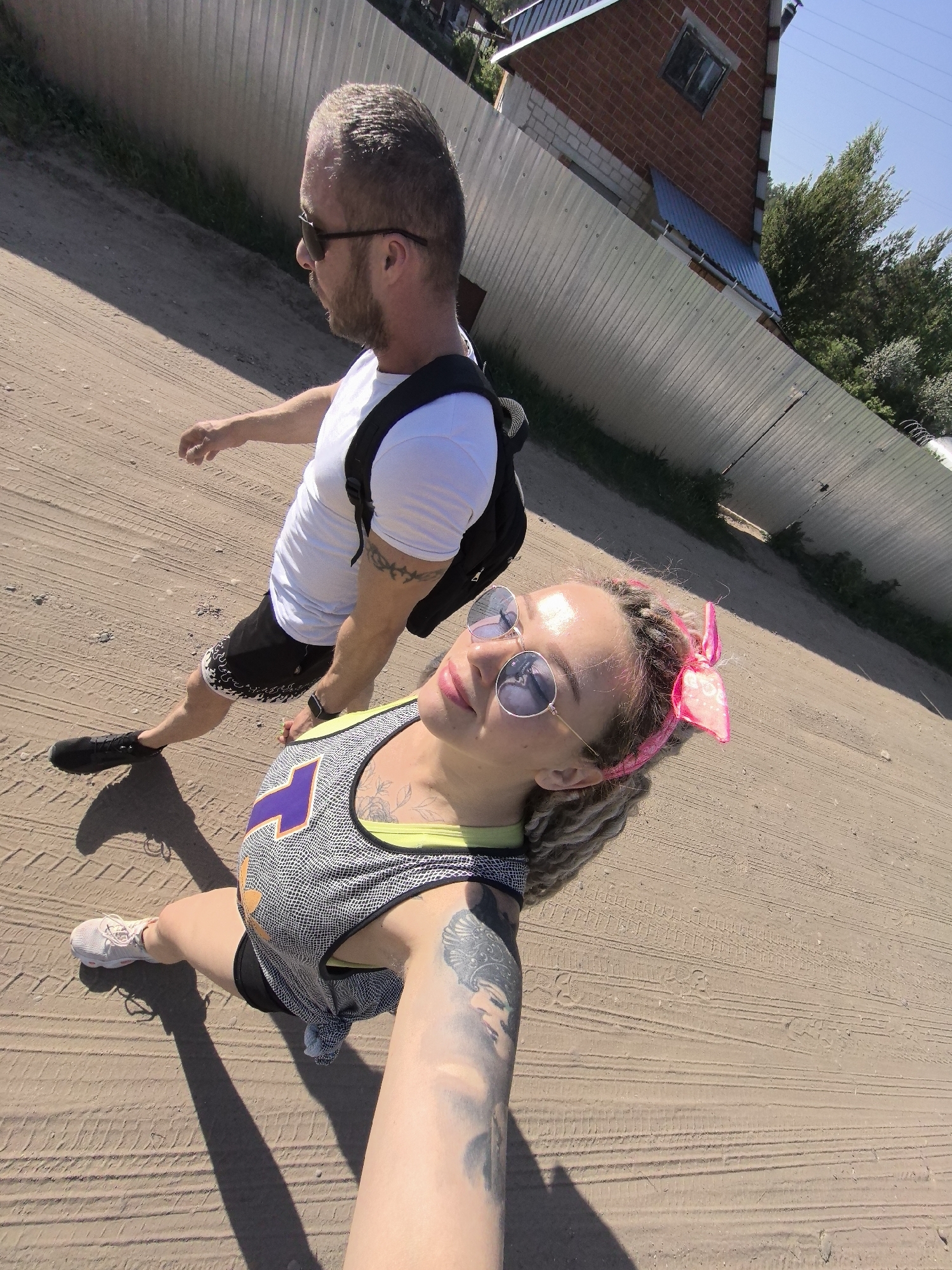 A medium-shot photograph captures a woman and a man walking on a dirt road in Noviy, Russia. The woman is in the foreground, looking up and left towards the camera. She is wearing sunglasses, a gray and white tank top with a purple number 7 on it, black shorts, and white sneakers. A pink bow is tied in her dreadlocked hair. Her left arm is extended toward the camera, revealing a large tattoo on her forearm. The man is behind her, wearing sunglasses, a white t-shirt, black shorts, black sneakers, and a black backpack. They are walking along a dirt road. Behind them is a silver fence with a glimpse of a brick house and some trees. The scene is brightly lit, casting shadows on the ground.