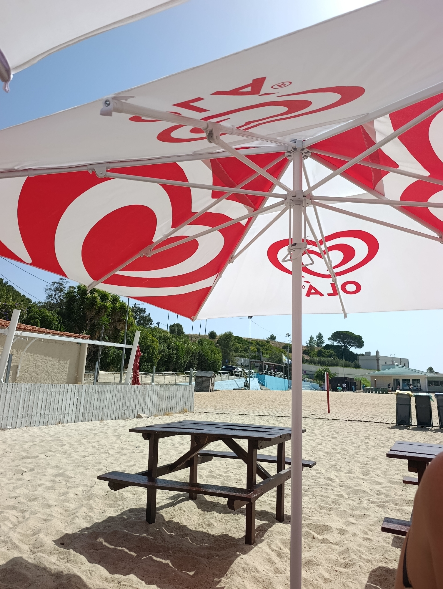 The image shows a sunny beach scene. In the foreground, a white umbrella with red logos (Ola brand) is casting a shadow on the sand. A wooden picnic table is centrally placed on the sand. In the background, there is a white wooden fence, some greenery, trees, and buildings. A swimming pool is visible to the left of the buildings. The sky is clear and blue. The scene appears to be at a beach resort or a public beach area in Setúbal, Portugal, given the presence of the Ola brand, which is a prominent ice cream brand in Portugal. There are no people clearly visible in the image, only blurred shapes that might be people in the distance. The environment is sandy and appears to be a recreational area.