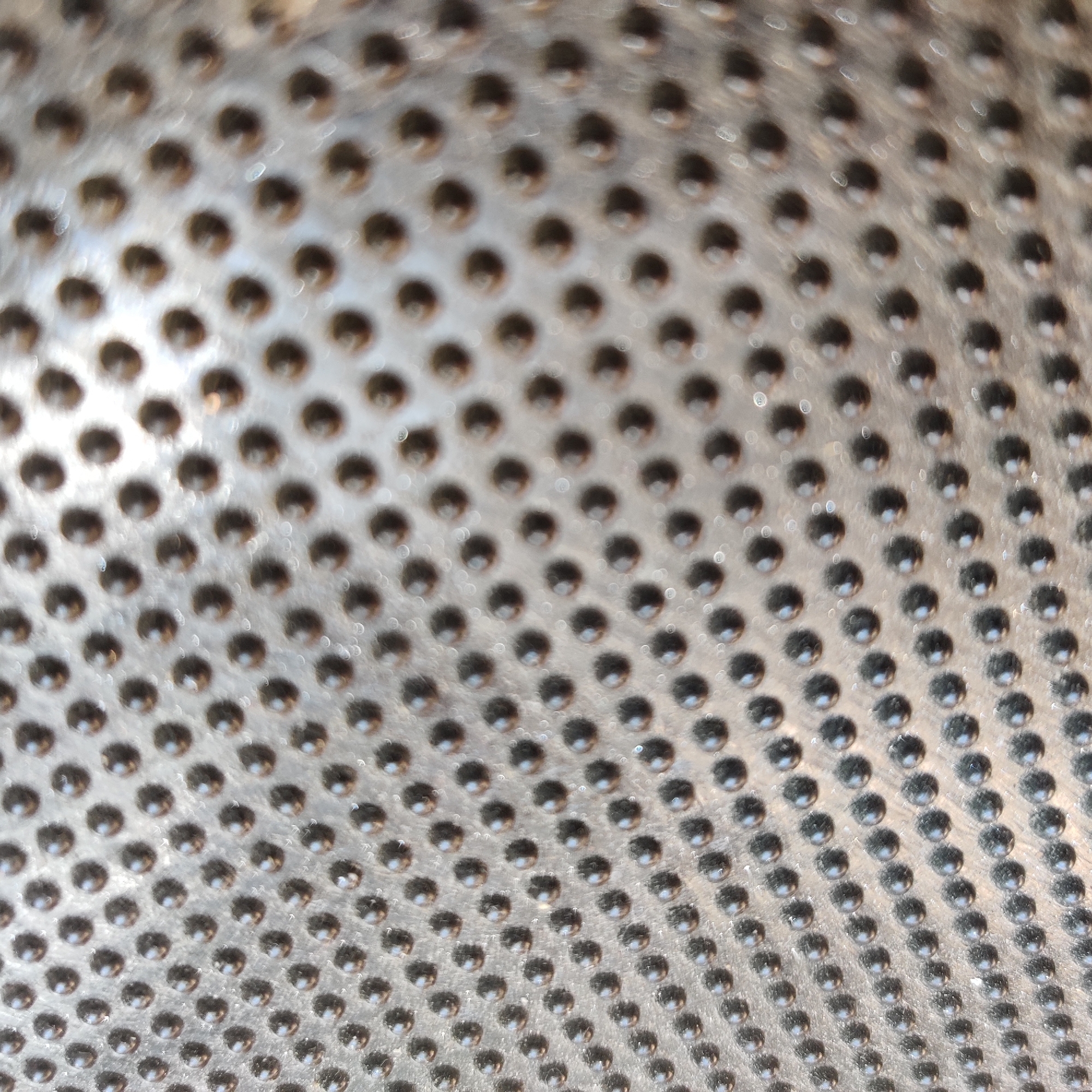 The image is a close-up, abstract shot of a textured metal surface. The surface is covered in a regular pattern of small, raised, dark, hemispherical bumps. The lighting creates highlights on the bumps and a bokeh effect in the background, suggesting the surface might be a floor mat or some other textured material. The overall impression is one of repetition and industrial design.
There is no discernible information in this image that can be used to determine a location in Yekaterinburg, Russia, or any other specific geographical location. The image is purely a macro view of a textured surface.