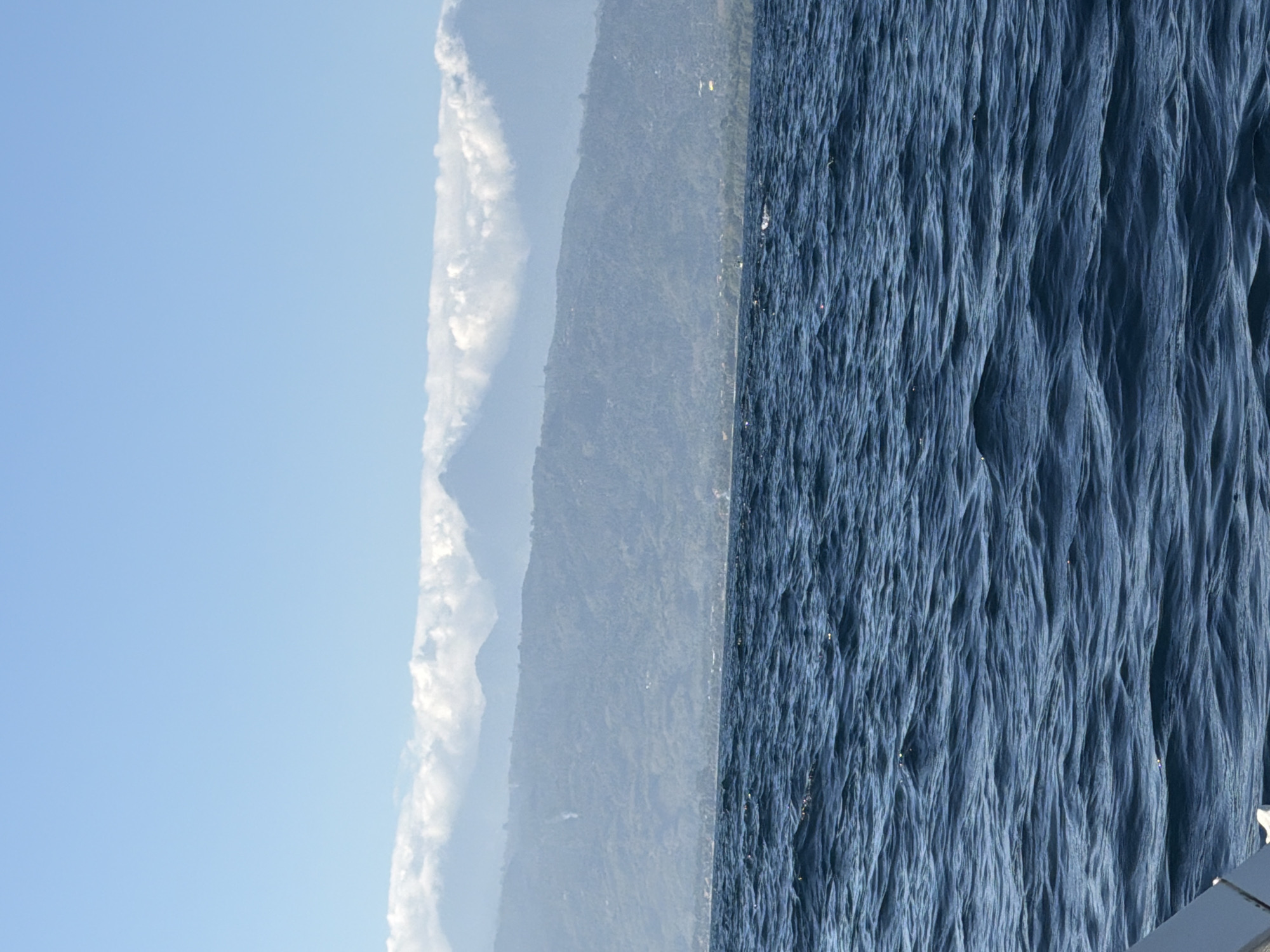 An image taken from a boat shows a view of a coastline with mountains in the background. The ocean in the foreground is a deep blue with gentle waves. The coastline features a strip of land with some vegetation and buildings, leading up to hazy, layered mountains. The tops of the mountains are partially obscured by clouds. The sky is a clear, light blue. The image appears to be taken during daylight, likely in the morning or afternoon, given the brightness and clarity of the sky. There is no indication of the specific location within Indonesia, other than it is along a coast.