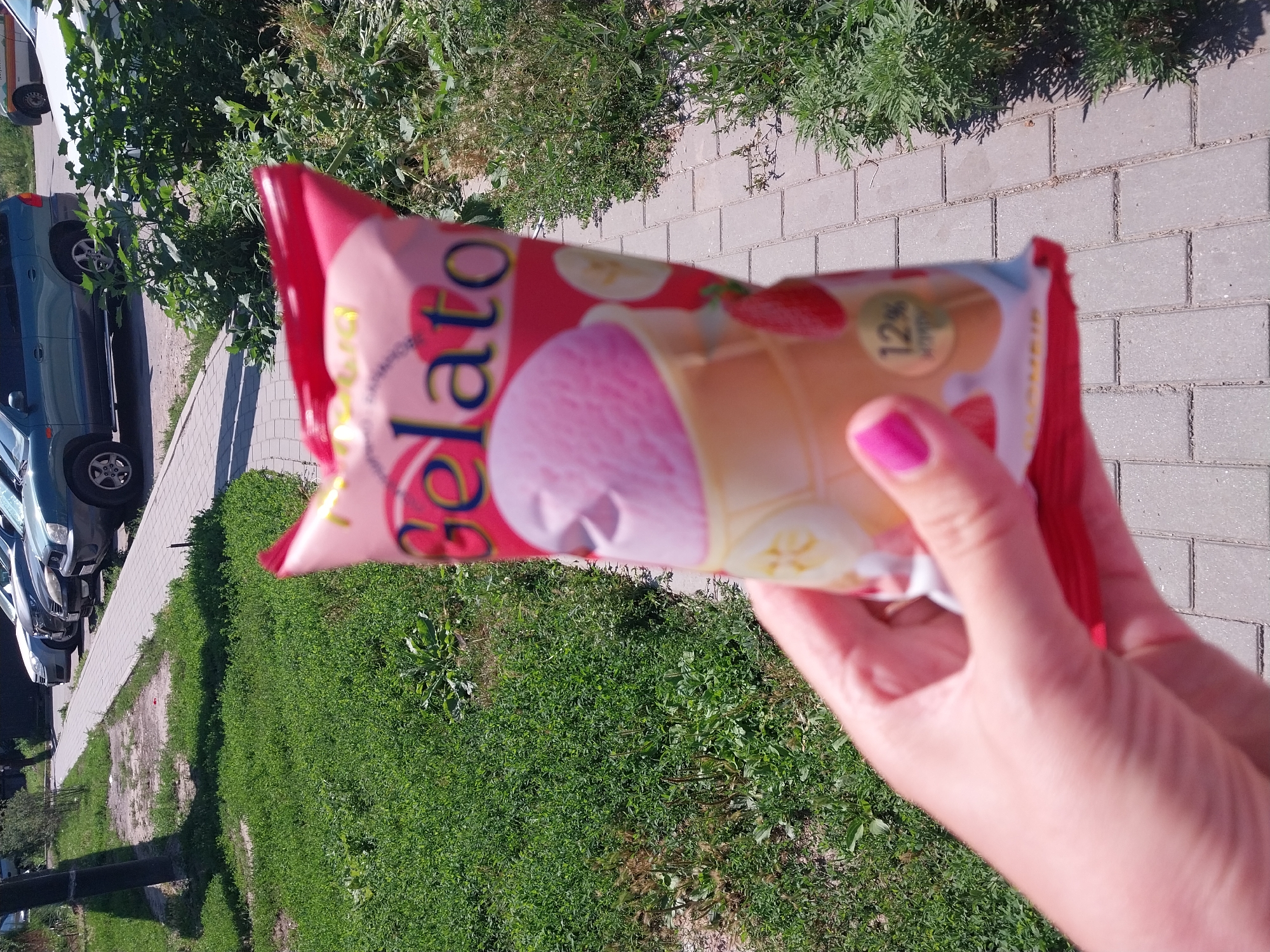 An individual's hand with bright pink fingernail polish holds a pink and yellow ice cream wrapper with the word "Gelato" prominently displayed in colorful lettering. The wrapper features images of strawberries and bananas. The background shows a paved walkway, green grass, and parked cars, suggesting an outdoor urban setting. This image was taken in Dnipro, Ukraine, on a sunny day.
