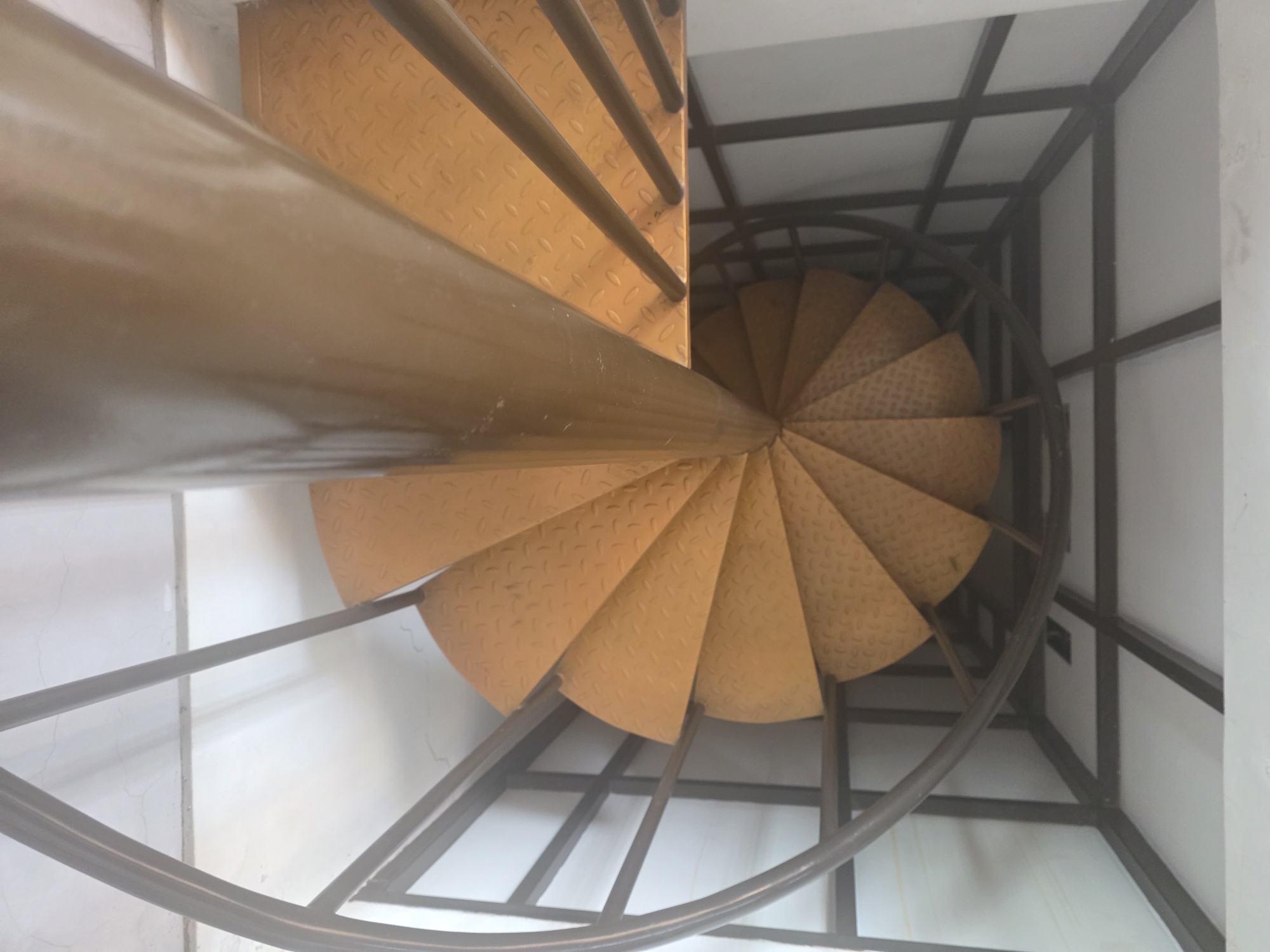 A top-down, close-up shot captures a spiral staircase. The staircase is constructed of metal, with wide, orange-colored steps featuring a diamond plate texture. A thick, cylindrical metal pole forms the central support. The staircase is enclosed by a dark metal frame, forming a square base. The surrounding environment appears to be an indoor space with white walls. The image is likely taken in Mekarsari, Indonesia.